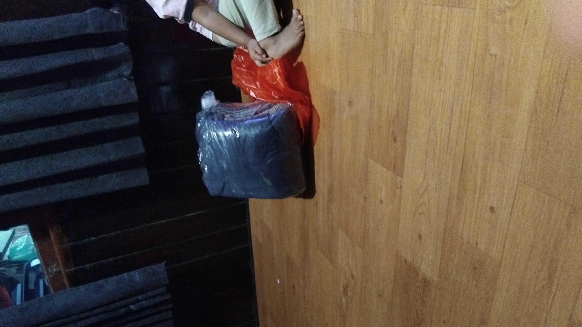 A person's bare feet and lower legs are visible on a wooden floor. To the left of the feet is a large, dark blue object wrapped in clear plastic, placed on top of a red plastic bag. The background shows a dark curtain and a wooden wall. The scene appears to be indoors, likely in a residential setting.