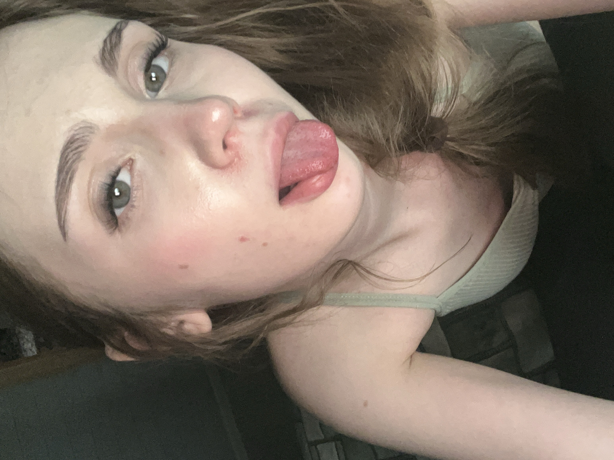 This is a close-up, slightly angled, indoor photograph of a young person. The subject's face is the primary focus, with their eyes open and looking towards the camera, and their tongue extended slightly from their mouth. Their eyebrows are well-defined and arched. The skin appears fair with some light blush on the cheeks and a few small moles. The hair is light brown and appears somewhat tousled.
The person is wearing a light-colored tank top with a ribbed texture. The background is dark and out of focus, with hints of what might be a textured surface and some vertical lines, possibly suggesting brickwork or paneling.
There is no visible text in the image, and no strong indicators of the time of day or weather. The overall mood conveyed is casual and perhaps playful due to the tongue gesture. The location context provided is Ashkadar, Russia, but there are no explicit visual cues in the image to confirm this.