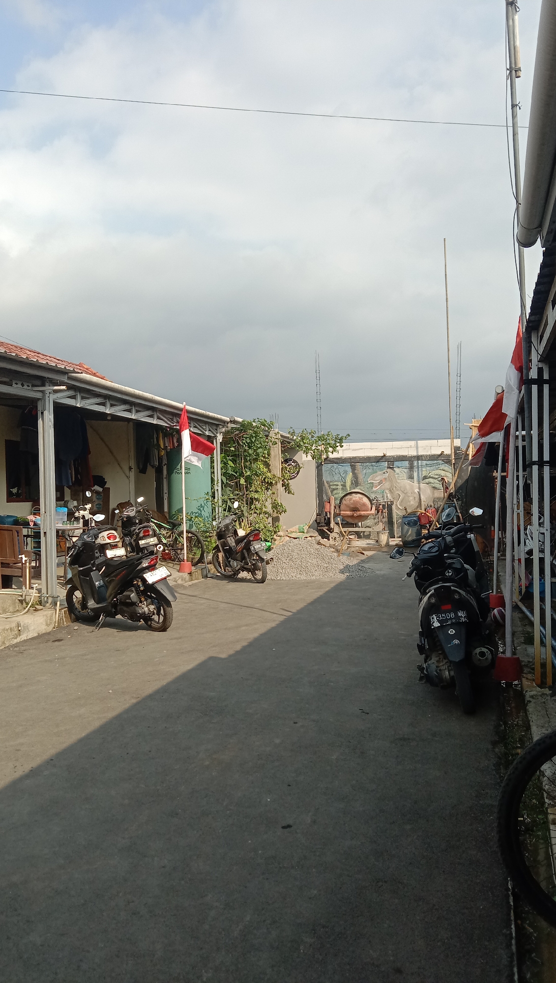 The image depicts an outdoor scene in Karangmangu, Indonesia, likely a residential or working area. The foreground is a paved pathway with shadows indicating sunlight from the left. Several motorcycles are parked along the pathway.
On the left side of the image, a building with an open-sided structure houses laundry hanging on a line and various objects. A red and white Indonesian flag is prominently displayed on a pole in front of this building.
Further into the scene, on the right side, there is a wall with a mural of a dinosaur. A concrete mixer and a pile of gravel are situated in front of the mural, suggesting ongoing construction or renovation. Another section of buildings or structures runs along the right edge, also adorned with red and white Indonesian flags. The sky is overcast with clouds.
There are no people visible in the image. The overall impression is one of a moderately occupied outdoor space with elements of daily life, construction, and national pride.