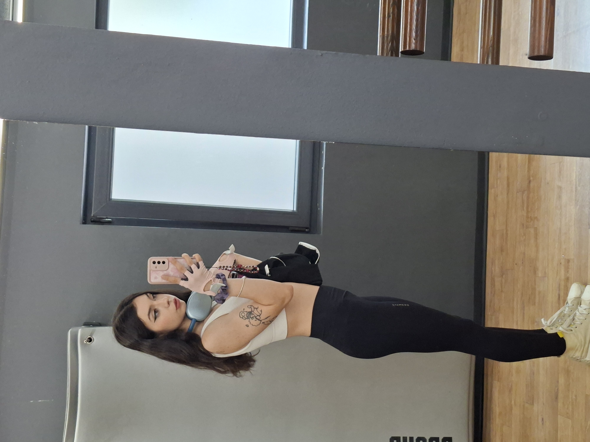 A young woman with long, dark hair is captured in a side profile, holding a smartphone to take a selfie in a gym. She is wearing a white sports bra, black leggings, and white high-top sneakers. Blue headphones are around her neck, and she has a tattoo on her left arm. She is also holding a black gym bag and pink wrist wraps. The background shows a gray wall, a large window, and wooden flooring, suggestive of an indoor fitness facility. The image was taken in Sofia, Bulgaria.