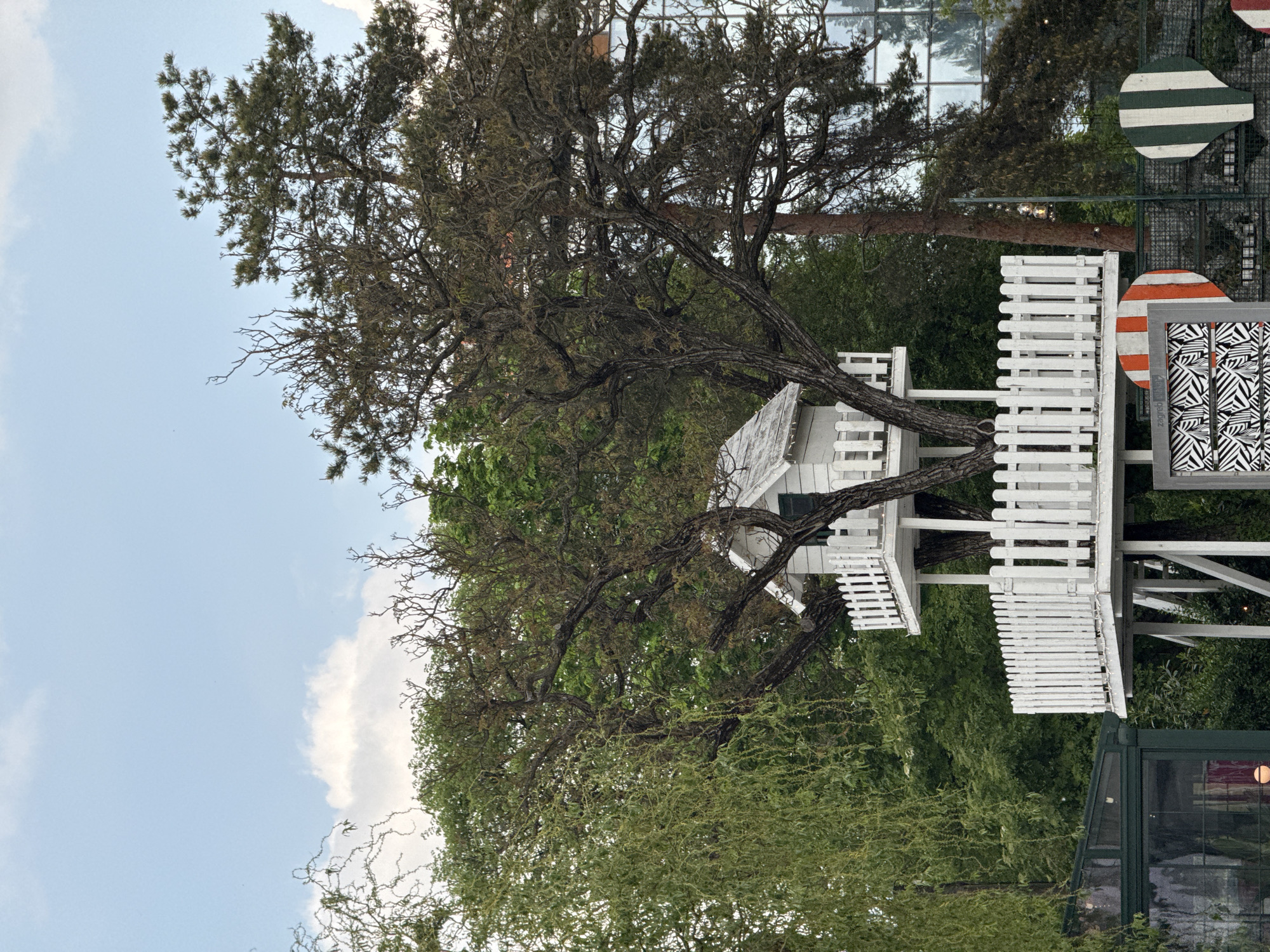 This image depicts a whimsical outdoor scene, likely situated in a park or garden in the City of Zagreb, Croatia. A prominent white treehouse is integrated into the branches of a large, mature tree, which appears to be a pine or similar conifer, characterized by its gnarled, dark branches and dense green-brown foliage. The treehouse features white wooden railings and blends with the natural structure of the tree.
The sky above is a clear light blue with soft white clouds, indicating it is daytime. Surrounding the treehouse are various other green trees and bushes, contributing to a lush environment. In the background, a modern building with large glass windows reflects the surroundings.
To the right of the treehouse, several decorative elements are visible within a metal grid framework. These include circular or oval shapes with distinct stripe patterns – one with green and white stripes, and another with orange and white stripes. Below these is a rectangular frame displaying a bold black and white zigzag design. On the side of this frame, the word "zagreb" is clearly written, with the letters "lly" or "lily" partially visible beneath it. In the bottom right corner, a structure with dark green frames and glass panels is partially visible. The scene is static, with no human activity taking place, showcasing a blend of natural beauty and artistic or architectural installations.