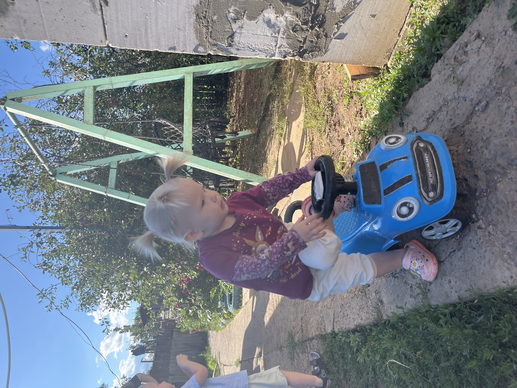 A young blonde girl with two pigtails, wearing a purple shirt with a deer design and white pants, sits in a blue toy car. She is holding the steering wheel and looking to her left. Her feet are in pink sandals. The car has black accents and "HZ SPEEDRUNNER" written on the side. The scene is outdoors, with a path made of concrete slabs and dirt, bordered by green grass and some leafy bushes. In the background, there is a green metal structure, a tree with red berries, and a blue sky with white clouds. Another child, wearing a light blue shirt and striped shorts, is visible in the lower part of the frame, with their back to the camera. The image appears to be taken in daylight, suggesting a sunny day. The setting looks like a backyard or outdoor play area in Rakhinka, Russia.