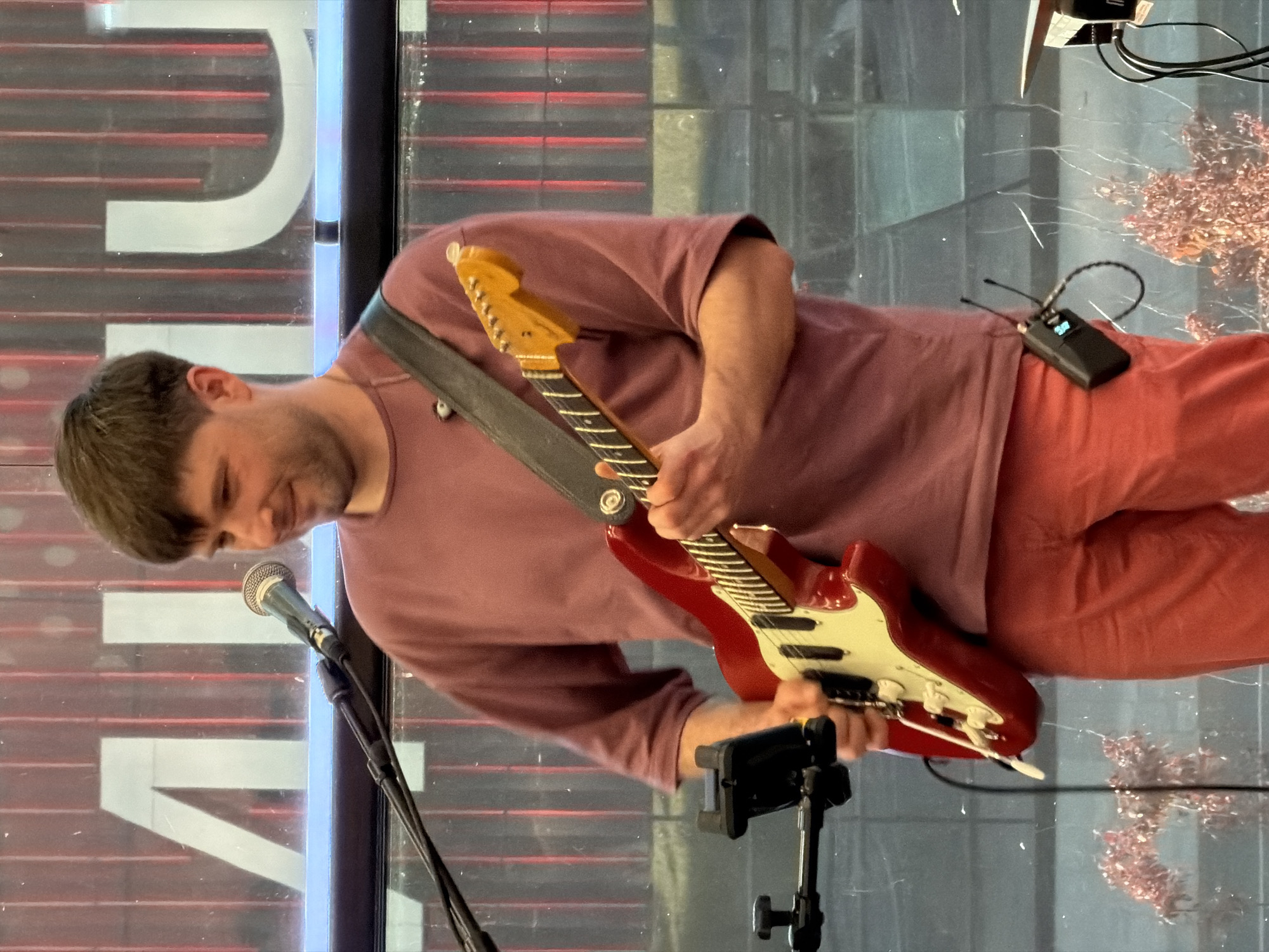 A fair-skinned man with dark hair plays an electric guitar, seemingly indoors near a large window with white geometric designs. He wears a muted pink long-sleeved shirt and similarly colored pants. The guitar is red with a white pickguard and a light-colored neck. A microphone stands on a metal stand to the left of the man. A wireless transmitter is clipped to his pants. A decorative element with a pinkish hue is visible to his right on a table. The city of Moscow, Russia, is not evident in the visual information provided.