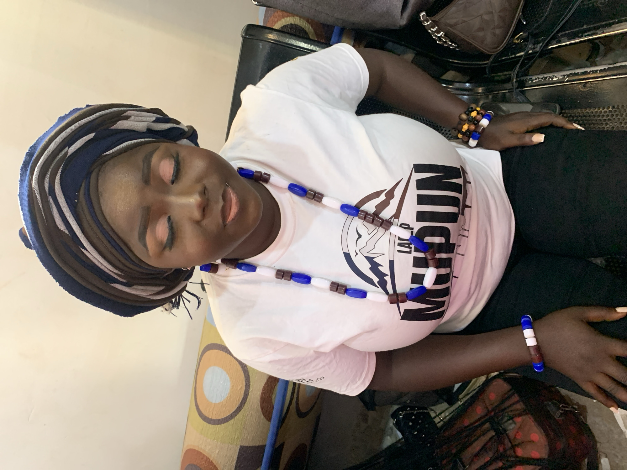A woman with dark skin sits indoors. She wears a striped blue, brown, and white head wrap, a white t-shirt with the word "MADUN" in black letters, a necklace with blue, brown, and white beads, and beaded bracelets on both wrists. She is wearing makeup and has manicured nails. She appears to be sitting on a cushioned chair or seat. Various items, including bags, are around her. The location is identified as Jos, Nigeria.