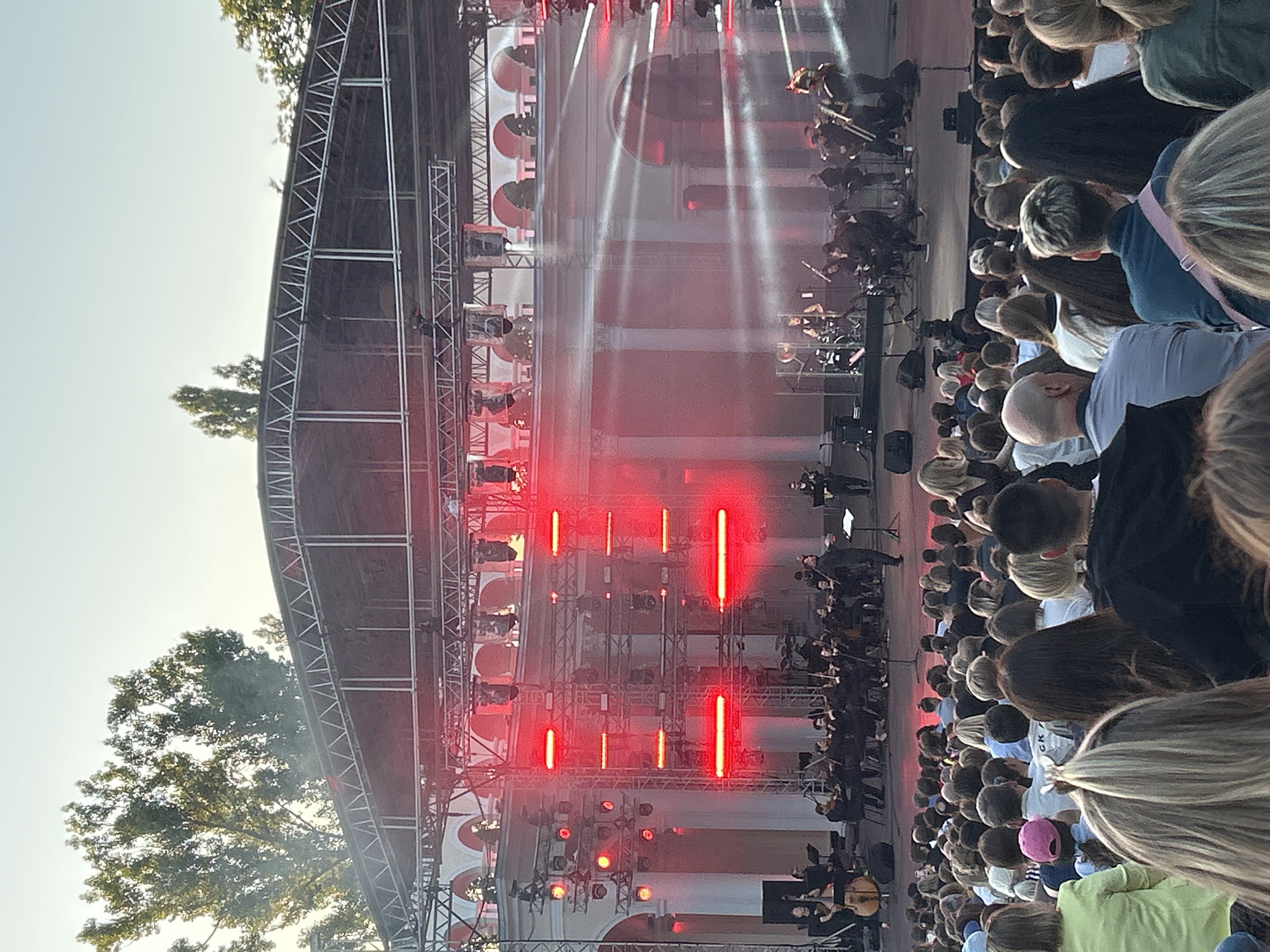 An outdoor concert is taking place at a venue resembling an amphitheater, possibly in Moscow, Russia. A large audience is seated, facing a stage set up with a complex lighting rig and scaffolding. An orchestra is performing on stage, with musicians playing various instruments, including strings and brass. A conductor is visible, leading the orchestra. The stage is illuminated with bright red lights, creating a dramatic effect. Behind the stage, a classical-style building with arches and columns is visible, suggesting a historical or culturally significant location. The sky above is clear, indicating it is likely daytime or early evening. The overall scene depicts a public cultural event with a performance for a gathered crowd.