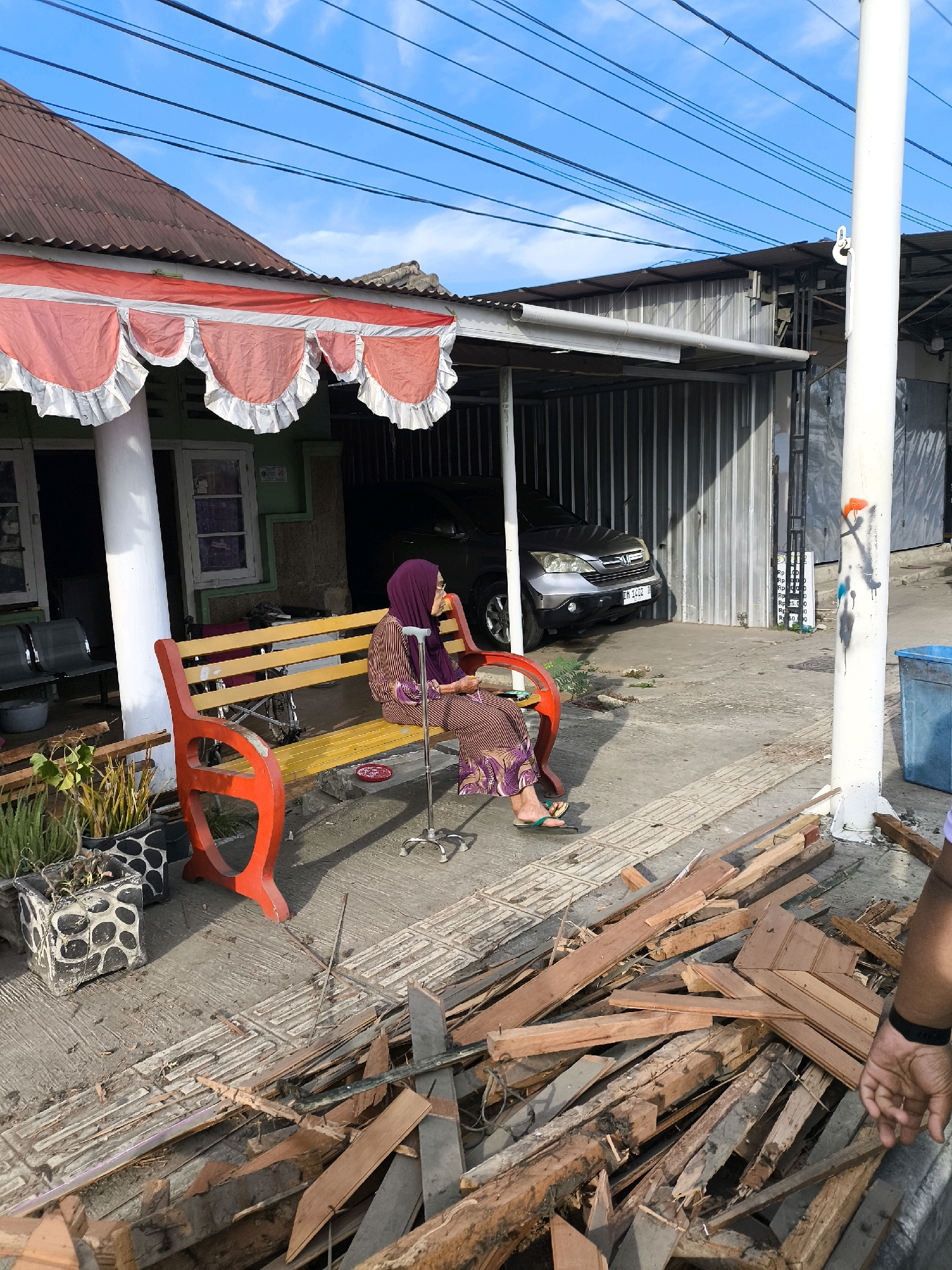 The image captures a street scene in Gorontalo, Indonesia. In the foreground, a pile of discarded wooden planks and debris lies on the concrete ground. To the left, a woman wearing a purple hijab and a patterned dress is seated on a yellow and red bench. She is holding a walking cane with a metallic base. Next to her on the bench is a wheelchair. In the background, a silver car is parked under a corrugated metal awning. A white utility pole stands to the right of the bench, adorned with graffiti. The sky above is blue with some scattered clouds, and power lines are visible. The environment appears to be a residential or commercial area with simple structures.
