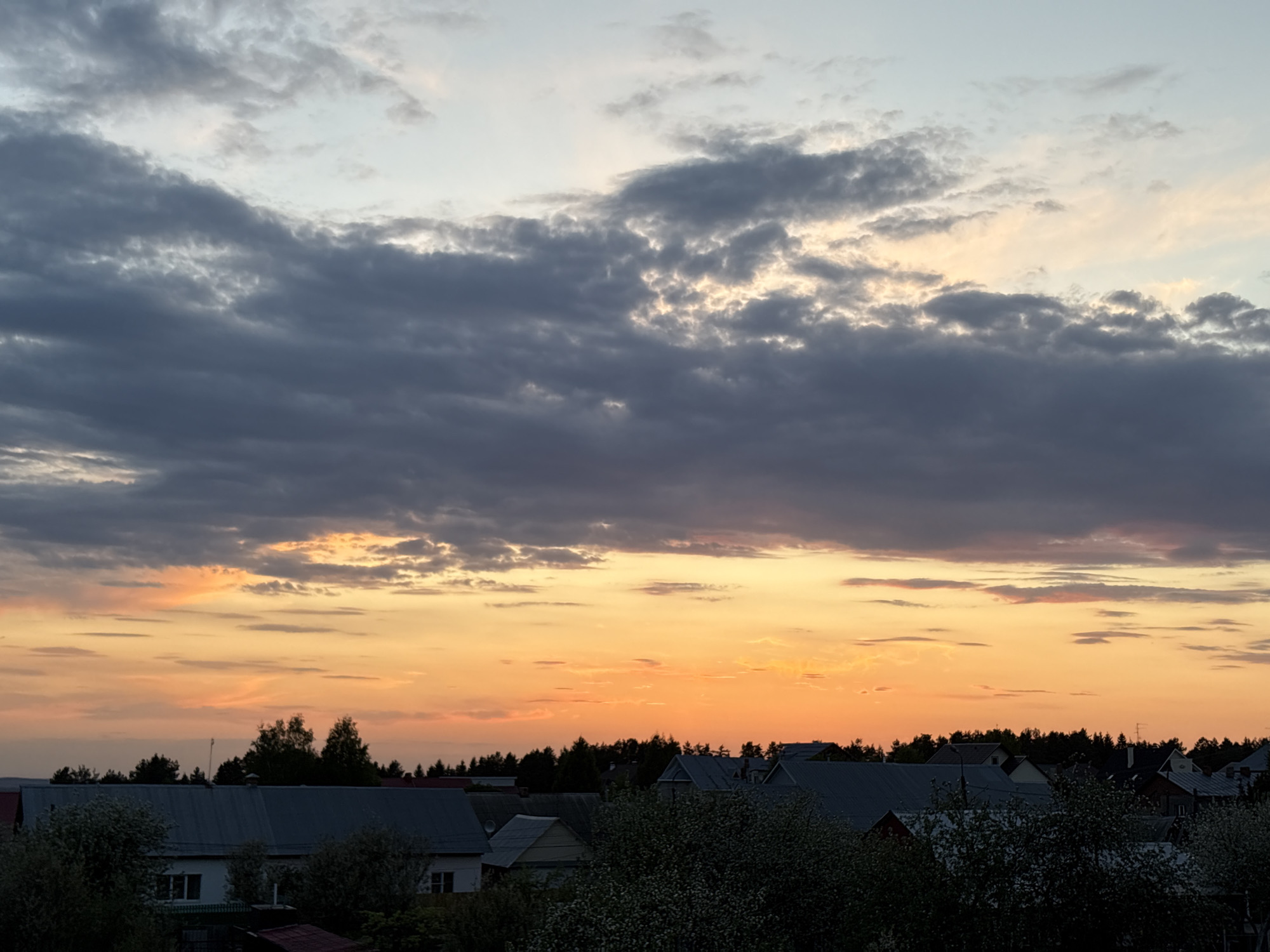 The image depicts a sunset scene over a residential area, likely in Прикамский, Russia, though specific identifiers are absent. The sky is a mix of colors, with dark, heavy clouds dominating the upper portion. Below the clouds, a band of orange and yellow indicates the setting sun. The foreground consists of the rooftops of houses and trees. The houses appear to be typical residential structures with pitched roofs. The trees are mostly dark silhouettes, with some displaying lighter foliage, possibly reflecting the sunset. No people or specific actions are visible in the scene.
