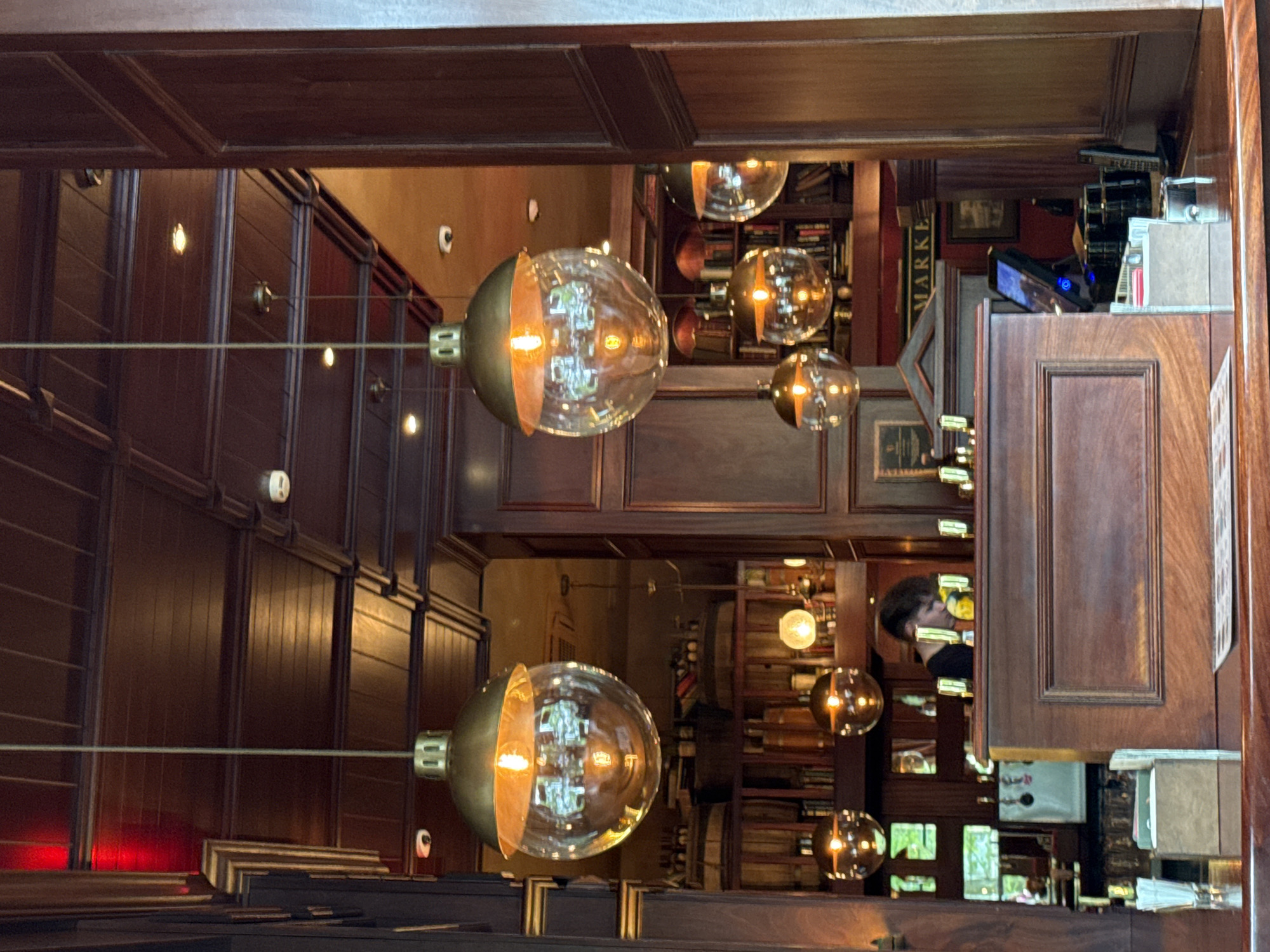 Here's a description of the image:
The image captures an interior scene with a warm and elegant aesthetic. The dominant features include unique light fixtures with spherical glass shades, accented by brass-colored metal components, which are suspended from the ceiling. The background suggests a library or a study, characterized by dark wooden shelves filled with books. The walls are also paneled with dark wood, contributing to the overall sophisticated ambiance.
In the upper right quadrant of the frame, there's a glimpse of what appears to be a reception or service area, with elements like a wooden counter and some visible equipment. A person is seen in the background, though their features are somewhat obscured.
The location is not determinable as Sochi, Russia, without more information or external reference points. The overall impression is that of a high-end establishment, possibly a private library, a club, or an upscale bar/restaurant.