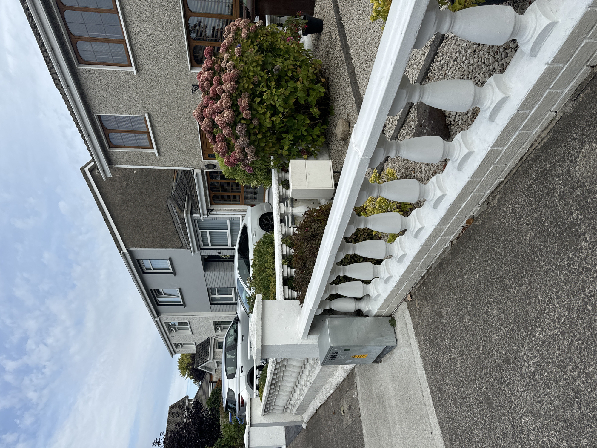 The image shows a residential street in Waterford, Ireland. The scene is focused on the front of several semi-detached houses, characterized by their grey rendered exteriors and tiled roofs. A prominent white balustrade with decorative pillars runs along the front of the properties, separating the pavement from the gardens.
In the foreground, a metal electrical box is visible next to the balustrade, with yellow hazard markings. The pavement is asphalt, and the garden areas are landscaped with gravel and a large hydrangea bush with pinkish-red blooms. Two cars are parked on the driveways: a silver hatchback and a white sedan. The sky above is cloudy.