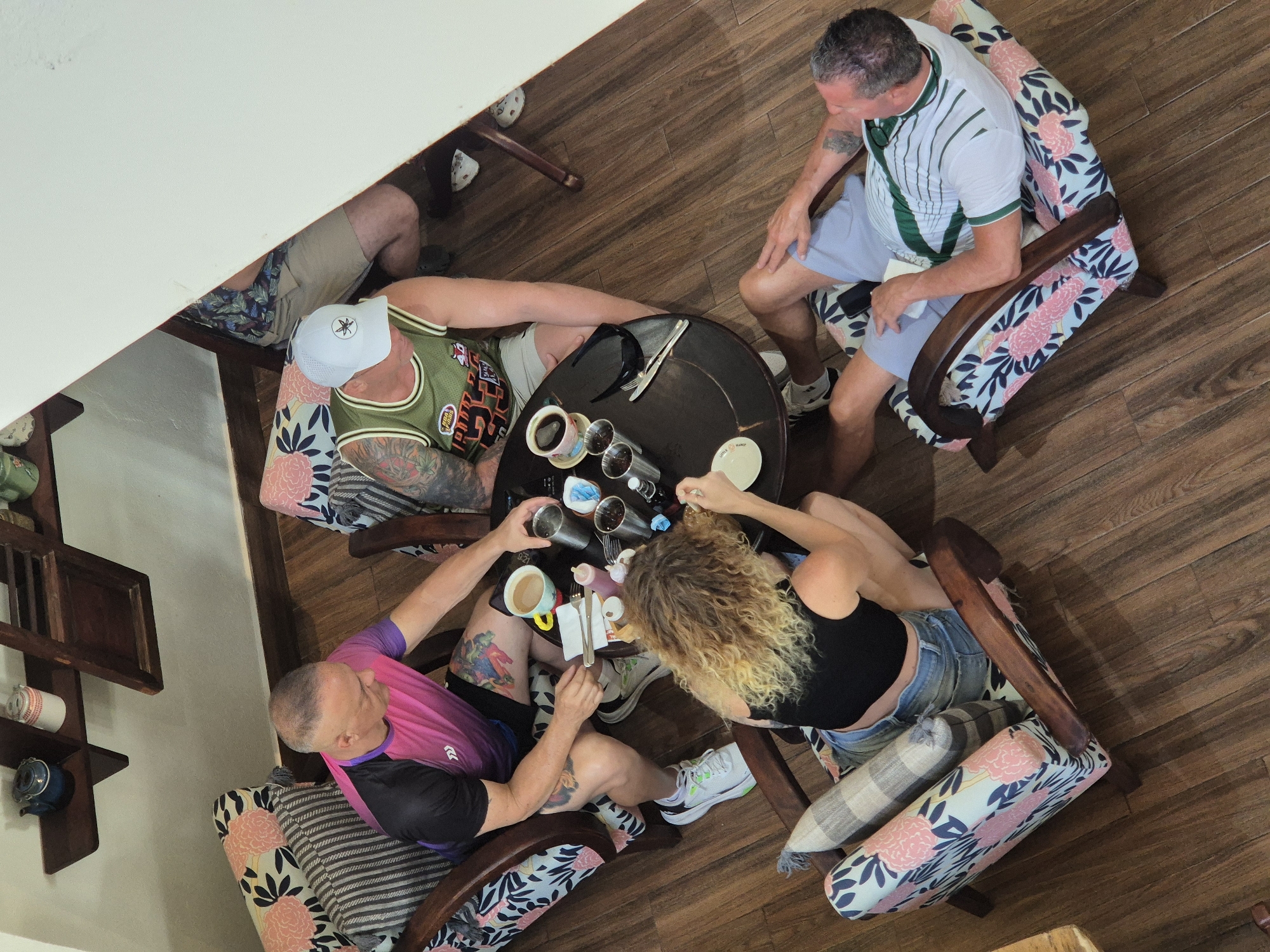 This image shows four individuals seated around a circular wooden table in what appears to be a cafe or restaurant. The setting is indoors, with wooden flooring and a white wall visible. A shelf unit with decorative items is mounted on the wall. The people are casually dressed and engaged in conversation or eating/drinking. On the table are coffee cups, drinks in metal containers, cutlery, and some condiments. The chairs are upholstered with floral and striped fabric. This scene is likely located in Đà Nẵng, Vietnam, given the provided context.