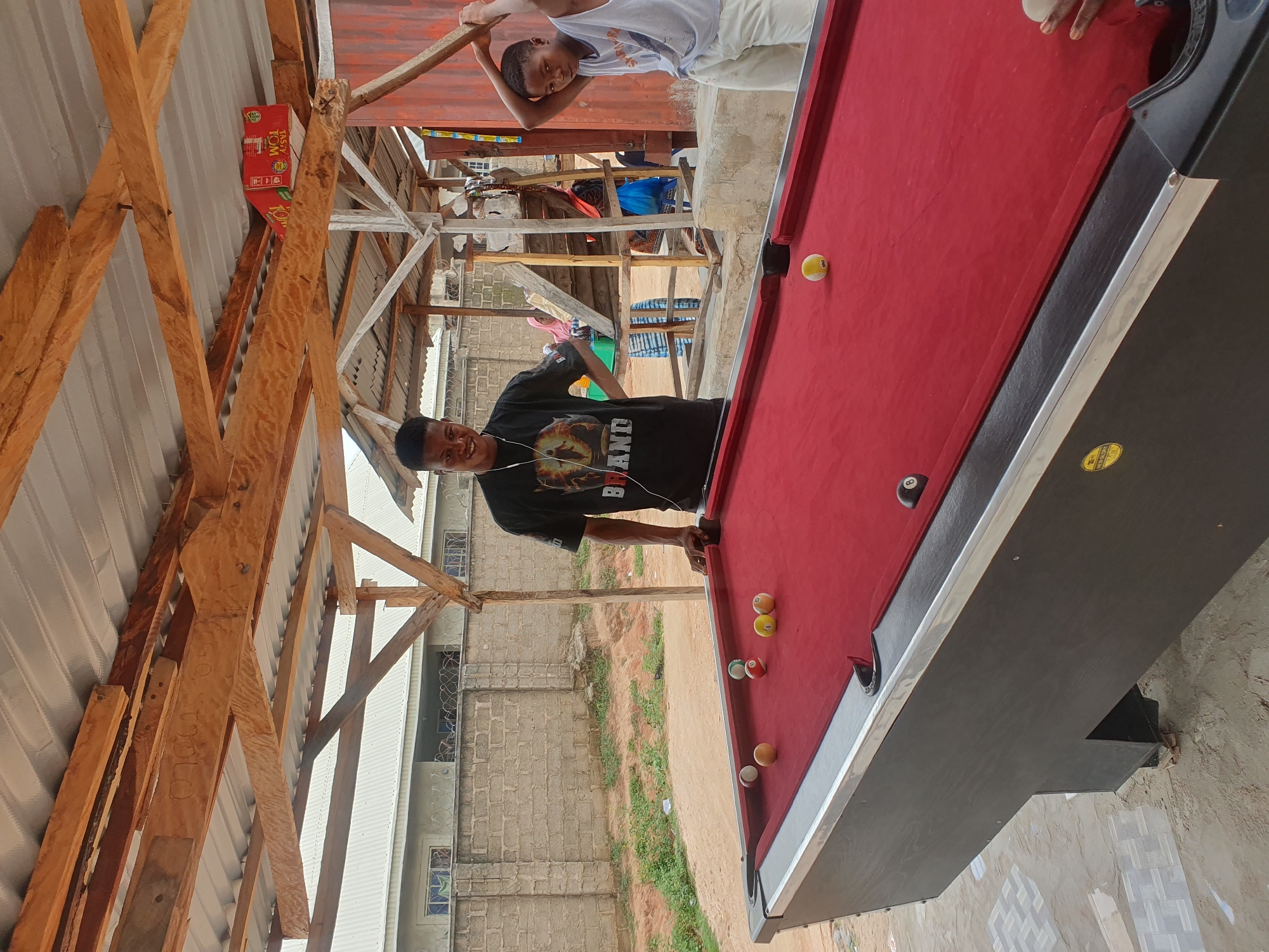The image depicts an outdoor pool table scene in Lokoja, Nigeria, likely on a sunny day. Two young males are the primary subjects. The one on the left, wearing a black t-shirt with graphics and earphones, is standing by the pool table, looking towards the viewer with a slight smile. The second male, on the right, is wearing a white tank top and light-colored shorts, holding a pool cue behind his head as if about to take a shot. Several pool balls are scattered on the red felt surface of the table, including a black 8-ball near the far pocket. In the background, a rough shelter with a corrugated metal roof supported by wooden beams is visible, suggesting a casual, open-air establishment. A brick wall and some greenery are also present behind the shelter, and a glimpse of a building with windows can be seen in the distance. The ground appears to be a mix of sand and dirt. The overall atmosphere is informal and suggests a recreational activity taking place.