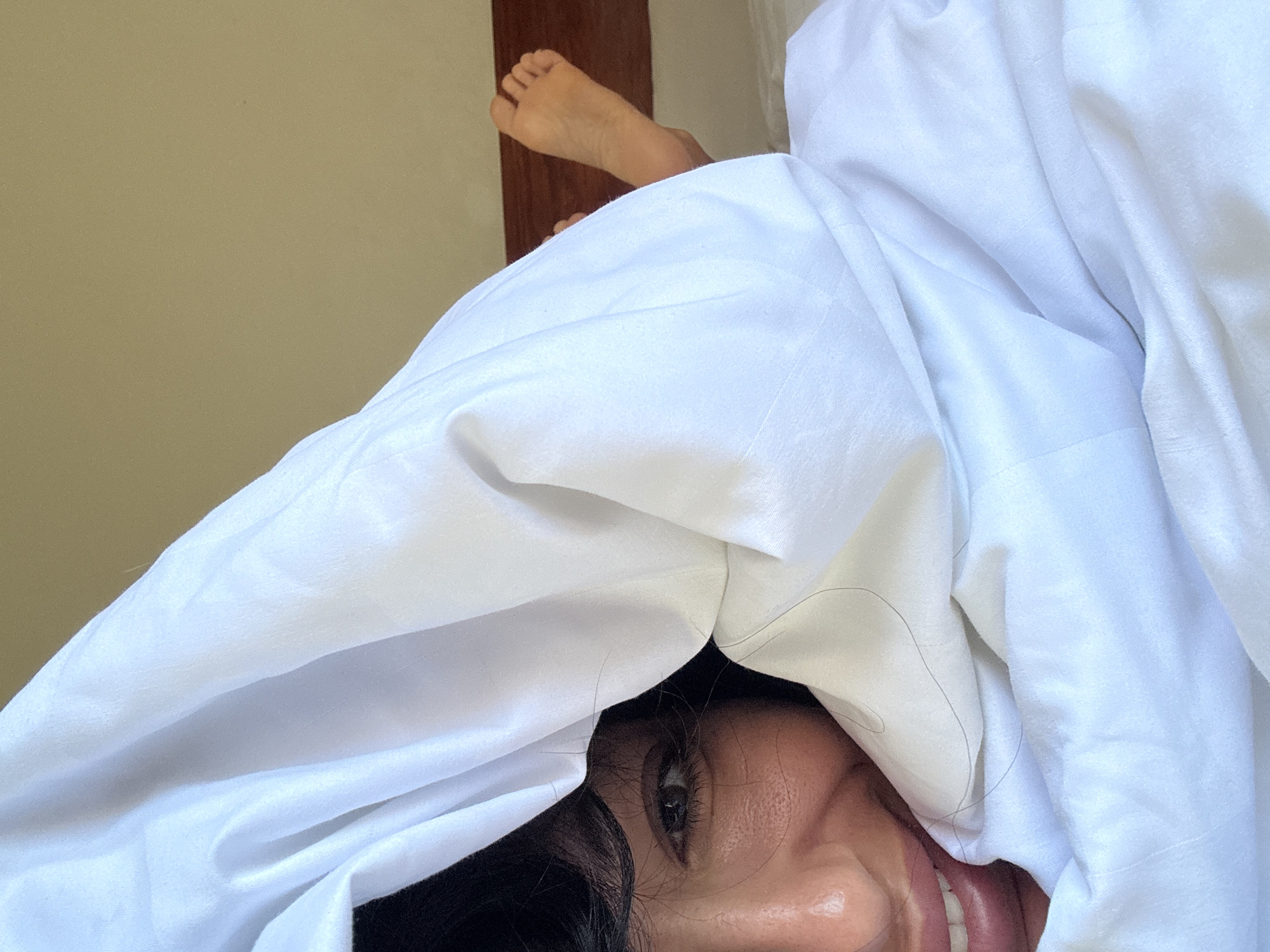 A woman is partially concealed under a white sheet, with only one eye and a portion of her smiling mouth visible. A pair of bare feet is also visible near a dark brown wall or doorframe. The image was taken in North Kuta, Indonesia.