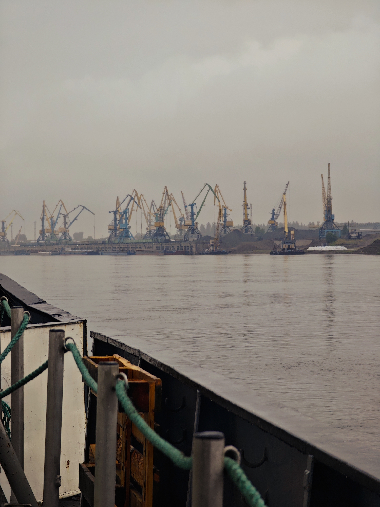 The image depicts a wide, overcast body of water, likely a river, in Lesosibirsk, Russia. In the background, a port facility is visible, characterized by numerous tall, industrial cranes. These cranes, painted in various colors including yellow, blue, and green, are arranged along a dockside. Piles of dark material, possibly coal or ore, are situated behind the cranes, indicating active cargo operations. The sky is a uniform grey, suggesting a cloudy or foggy day, which contributes to the muted tones of the scene. In the foreground, the edge of a vessel is visible, with a dark railing and some nautical rigging, including green ropes threaded through metal posts. The perspective suggests the photograph was taken from another vessel on the water, looking towards the port. There are no visible people in the image.