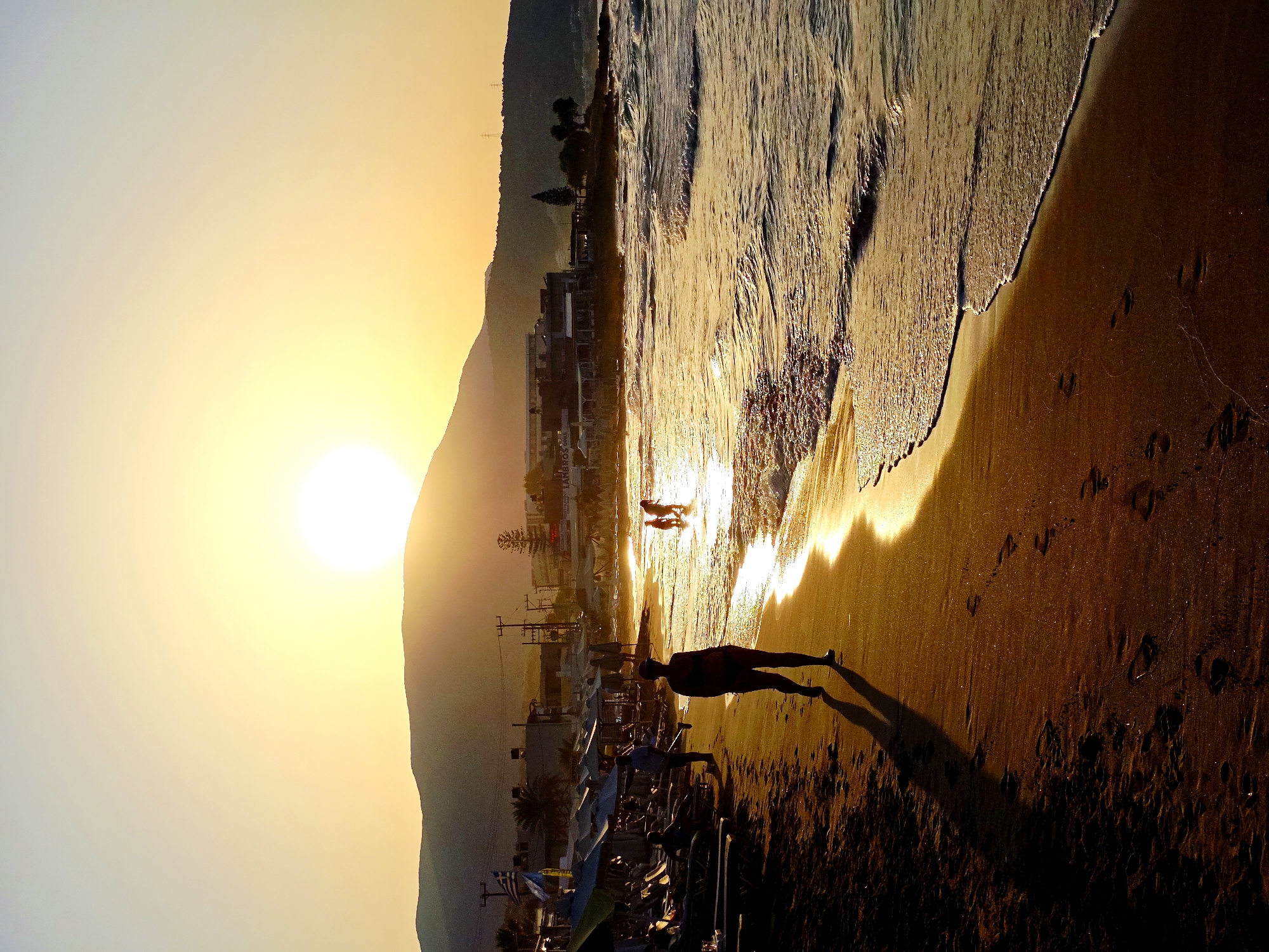 A sunset scene on a beach in Malia Municipal Unit, Greece, is captured. The sun is low on the horizon, casting a warm golden glow across the sky and reflecting on the water. A large, hazy sun is visible above a dark, silhouetted mountain range in the background.
In the foreground, a sandy beach stretches along the coastline. The wet sand near the water's edge is illuminated by the setting sun, and footprints are visible. On the left side of the beach, beach chairs and umbrellas are set up, with a few people partially visible in silhouette. A lone figure walks along the shoreline in the mid-ground, their long shadow stretching out before them.
The sea is calm, with gentle waves rolling towards the shore. The water reflects the warm colors of the sunset, creating a shimmering effect. In the distance, several structures that appear to be buildings or tavernas are visible along the coastline, also silhouetted against the bright sky. A few palm trees and other vegetation dot the landscape. Two small figures are seen wading in the shallow water near the shoreline in the mid-ground. The overall impression is one of a tranquil evening at the beach.