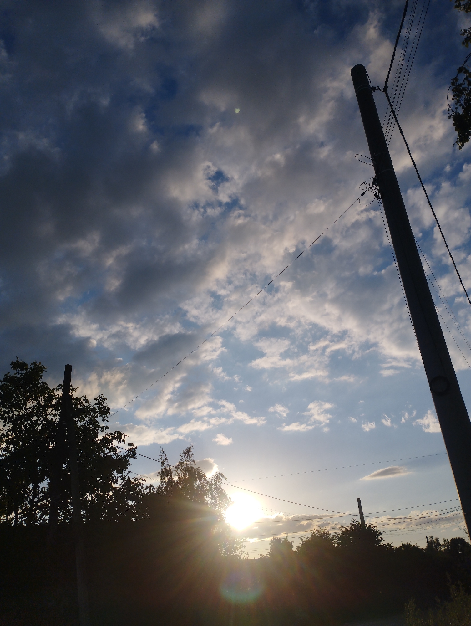 A low-angle, full shot captures a cloudy sky at sunset above a silhouetted treeline and power poles. The sky is filled with dramatic, dark clouds interspersed with patches of bright blue and illuminated by the sun which is low on the horizon, creating bright lens flares and radiating sunbeams. Two tall, dark utility poles with numerous wires are visible, one dominating the right side of the frame and another shorter one partially obscured by trees on the left. The foreground is mostly dark, showing the silhouettes of trees and dense foliage. The location is listed as Unknown, Unknown.