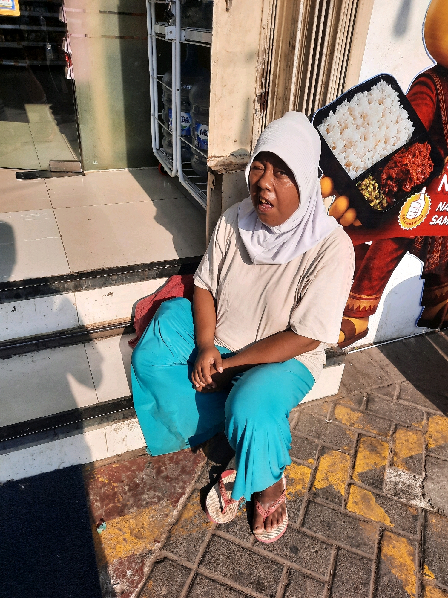 An individual wearing a light-colored headscarf and a plain t-shirt is seated on the steps of a building, with their legs extended onto the tiled pavement outside. They are also wearing bright blue trousers and pink flip-flops. The person's mouth is open, and they appear to be looking slightly upwards and to the left. Behind them, a large advertisement featuring a meal with rice and side dishes is visible. To the left of the image, a store entrance with glass doors is present, and a rack with several large water jugs is situated near the building's entrance. The ground is paved with grey tiles and sections of red and yellow painted markings. The scene appears to be outdoors, in a commercial area within Babakan Madang, Indonesia, as indicated by the presence of the store and the advertisement.