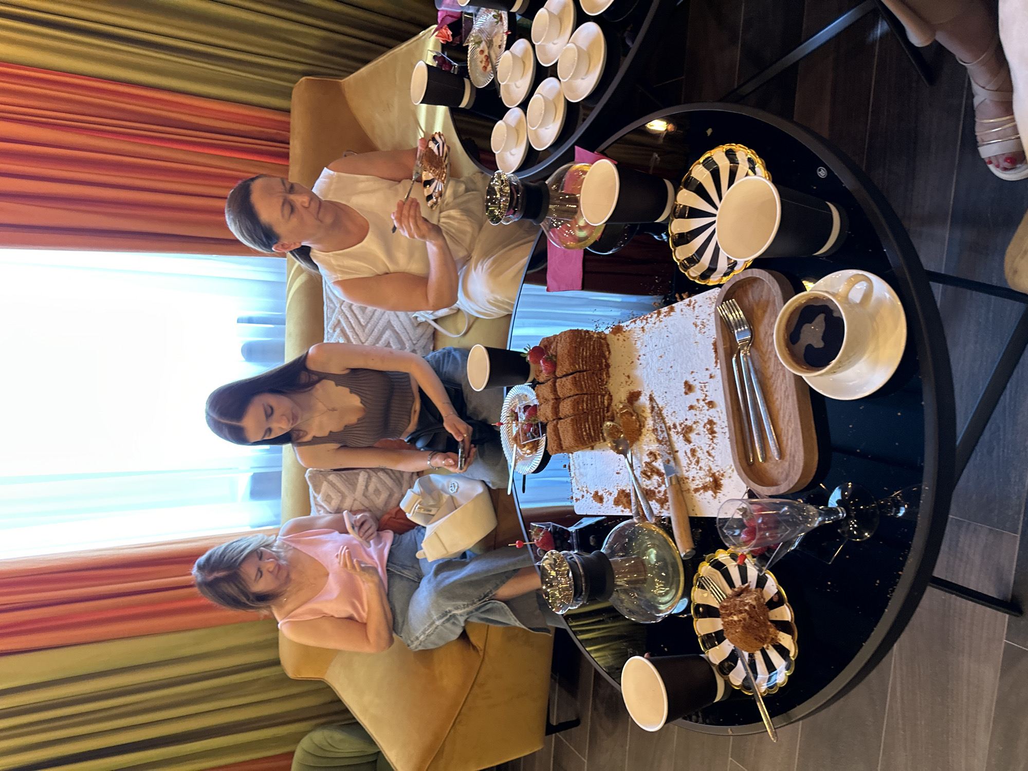 Three women are seated on a yellow sofa, attending to their mobile phones or eating cake. In the foreground, a round black table holds a variety of items, including a cake, coffee cups, plates with cake slices, and a glass of champagne with berries. The background features colorful curtains and a window. The scene appears to be set indoors, possibly in a cafe or a private residence in Sochi, Russia.