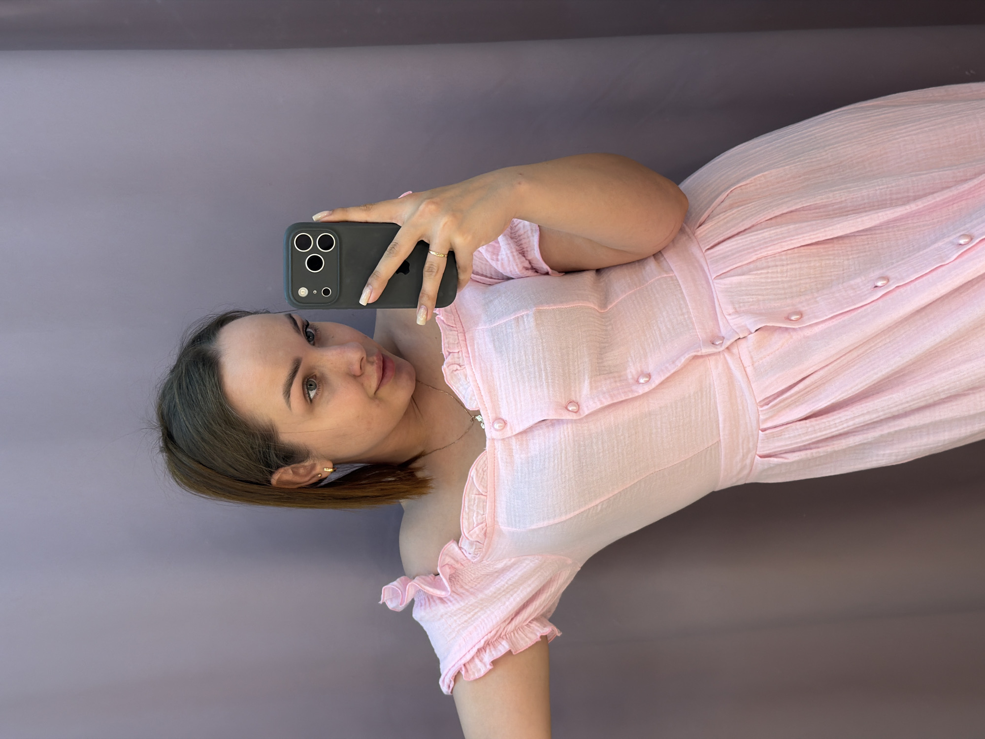 A woman is taking a selfie with a black-cased smartphone. She has short, brown hair and is wearing a pink dress with ruffles on the sleeves and buttons down the front. She is wearing a ring on her finger and a necklace. The background is a plain, light brown color. The picture does not provide enough information to determine the location in Chaikovsky, Russia, or any further details about the environment.