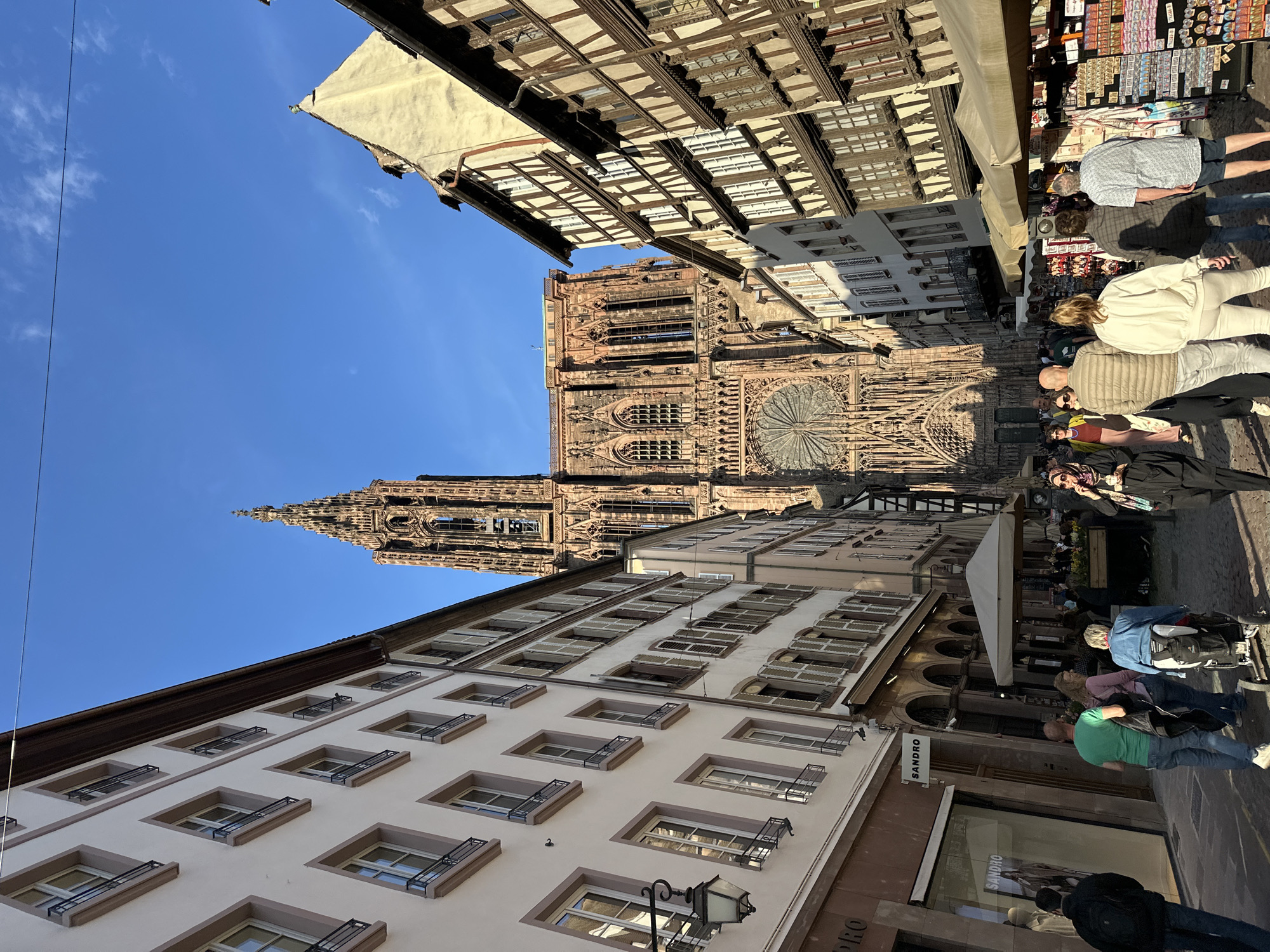 The image captures a street scene in Strasbourg, France, dominated by the imposing Strasbourg Cathedral in the background. The cathedral's intricate facade and spire are prominently displayed. Flanking the street are buildings with varying architectural styles, including traditional half-timbered structures and buildings with multiple windows and balconies. People are walking along the street, some appearing to be tourists browsing stalls selling souvenirs. The sky is clear and blue. The image gives the impression of a vibrant urban environment.