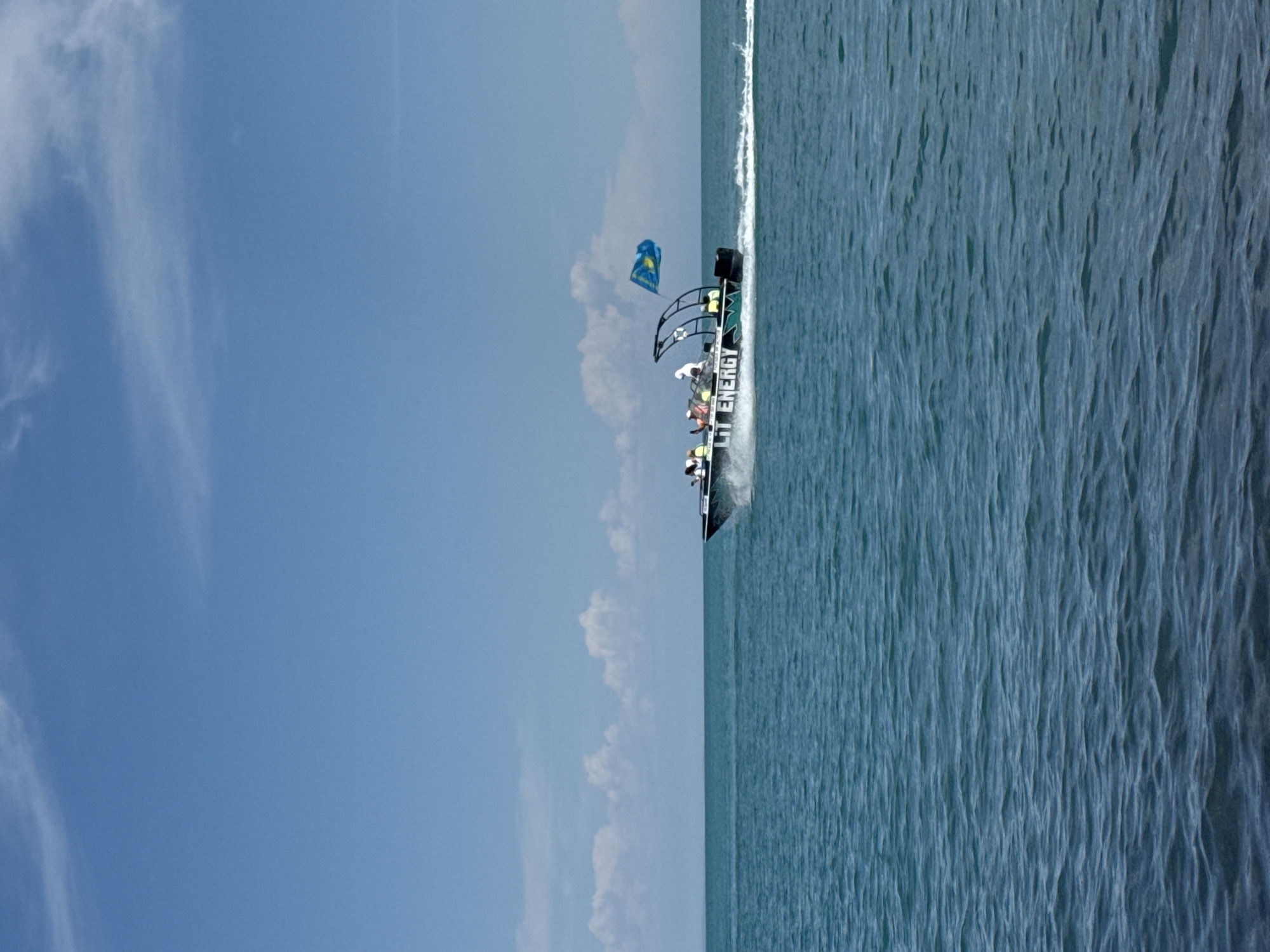 A speedboat with the words "LIT ENERGY" emblazoned on its side is moving across the water, creating a wake. Several people are visible on the boat, some standing and some seated. A blue and yellow flag is attached to a pole on the boat. The scene appears to be taking place on a body of water with a clear blue sky above, dotted with a few white clouds. The location is specified as the city of Ақши, Kazakhstan.