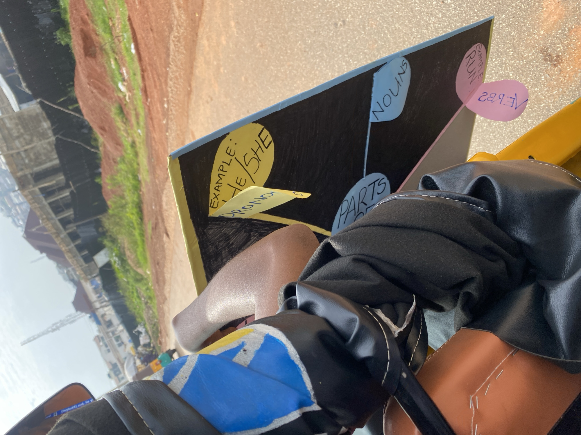 The image shows a close-up view of a triangular blackboard with handwritten notes on it, likely related to grammar lessons. The blackboard is held by a person whose arm and shoulder are visible, wearing black fabric. The background suggests an outdoor urban environment with buildings under construction, red dirt mounds, and vegetation. The overall scene is captured from a low angle, possibly from inside a vehicle.
The blackboard has sections labeled "EXAMPLE: HE/SHE" and "PRONOUN". Another section is labeled "PARTS" and there's a label "NOUNS". A pink sticky note with writing on it is attached to the bottom of the blackboard. The writing on the sticky note is difficult to decipher but appears to be in English. The person holding the blackboard is wearing a black garment with white stitching. The background indicates a sunny day with a cloudy sky. The location is identified as Rafin Par, Nigeria, which is inferred from previous context and not directly visible in the image.