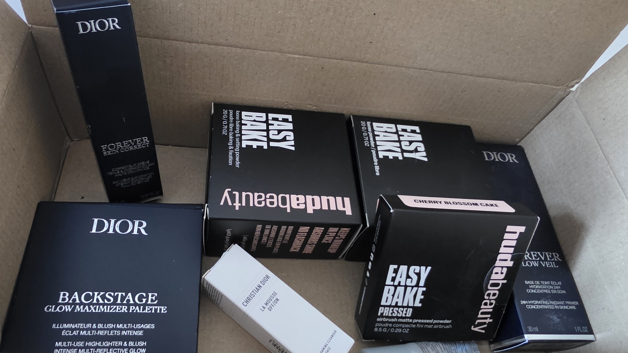 The image displays the interior of an open, brown corrugated cardboard box filled with various new, boxed cosmetic products. The contents primarily consist of items from the brands Dior and Huda Beauty. Visible Dior products include a "FOREVER SKIN CORRECT" bottle/box, a "BACKSTAGE GLOW MAXIMIZER PALETTE" box, and a smaller white box for "CHRISTIAN DIOR LA MOUSSE OFF/ON." Partially visible is another tall black Dior box for "FOREVER GLOW VEIL." Huda Beauty products include multiple black boxes of "EASY BAKE" loose baking & setting powder (20 G / 0.71OZ) and one "EASY BAKE PRESSED" airbrush matte pressed powder box (8.5 G / 0.29 OZ) with a "CHERRY BLOSSOM CAKE" label. The items are arranged loosely within the box, some upright and some oriented horizontally, suggesting storage, packing, or unpacking. No individuals, actions, or specific interactions are depicted. The environment is confined to the interior of the box. The precise location within the city of London, United Kingdom, cannot be determined from the visual information provided.