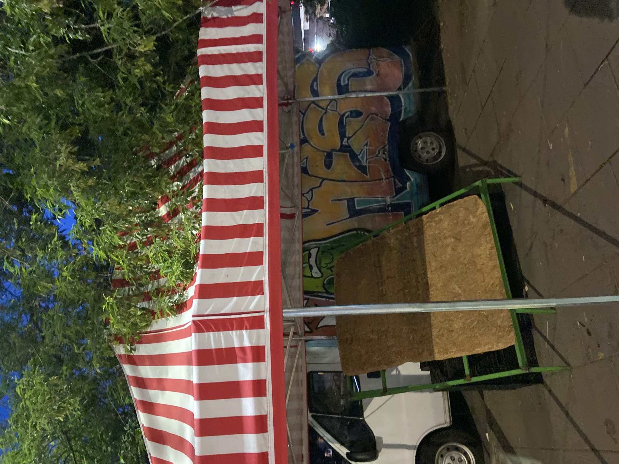 The image shows a market stall with a red and white striped canopy. Beneath the canopy, a white van is visible, partially covered by graffiti. The van appears to be parked on a street or sidewalk, with trees and foliage overhead. A green metal stand holding a piece of wood is positioned in front of the van. The scene is dimly lit, suggesting it is nighttime or evening. The image does not provide any specific details that would allow for identification of its exact location within Warsaw, Poland.