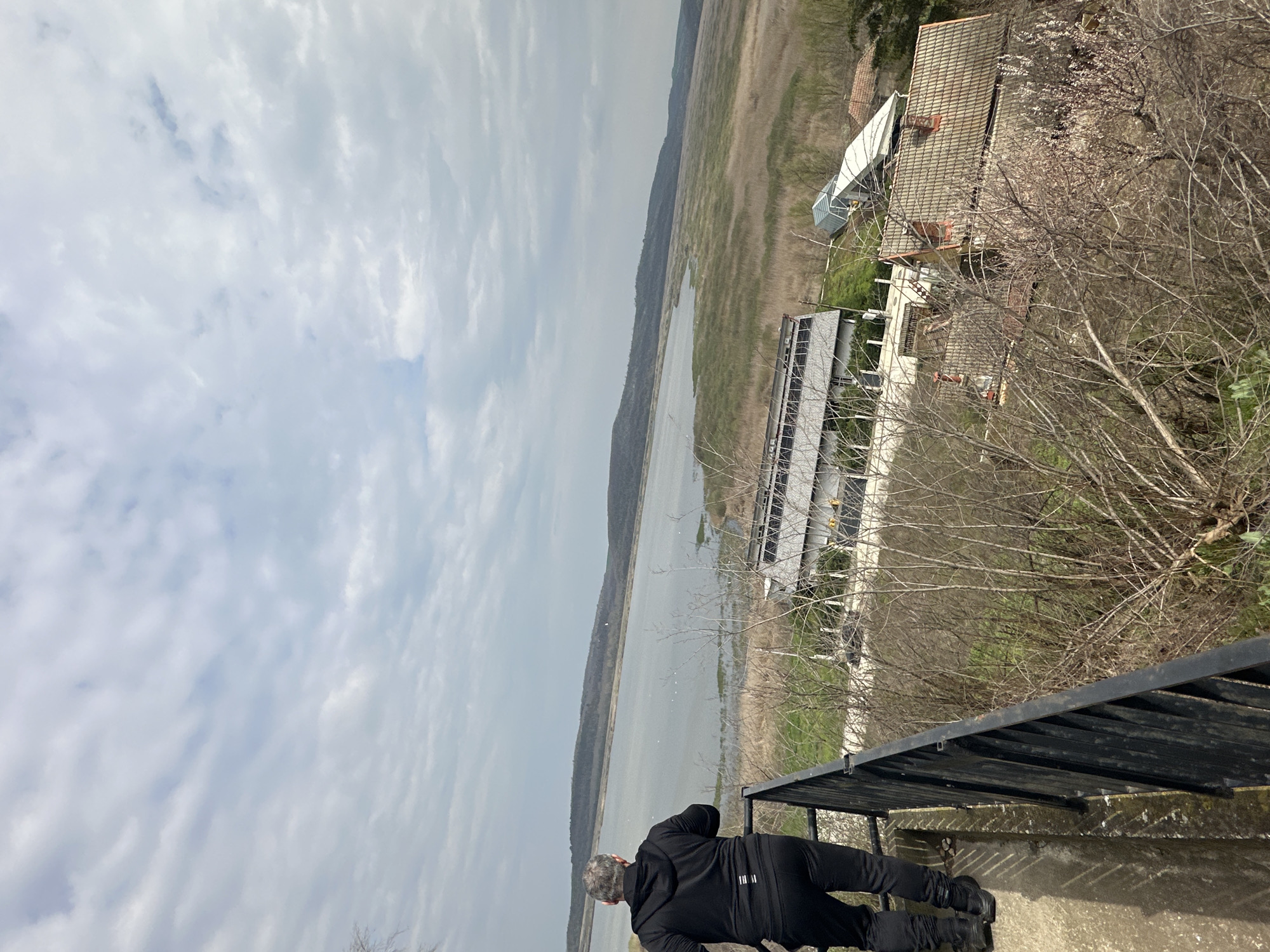 A person dressed in black is observing a landscape. The individual is positioned on a concrete structure with a black metal railing. In the distance is a body of water bordered by grassland and trees. To the right of the body of water is a building with a tiled roof and a smaller white structure. A multi-story building is located behind the railing and a leafless tree. The sky is overcast with clouds. This scene depicts the Srebarna Nature Reserve in Bulgaria.