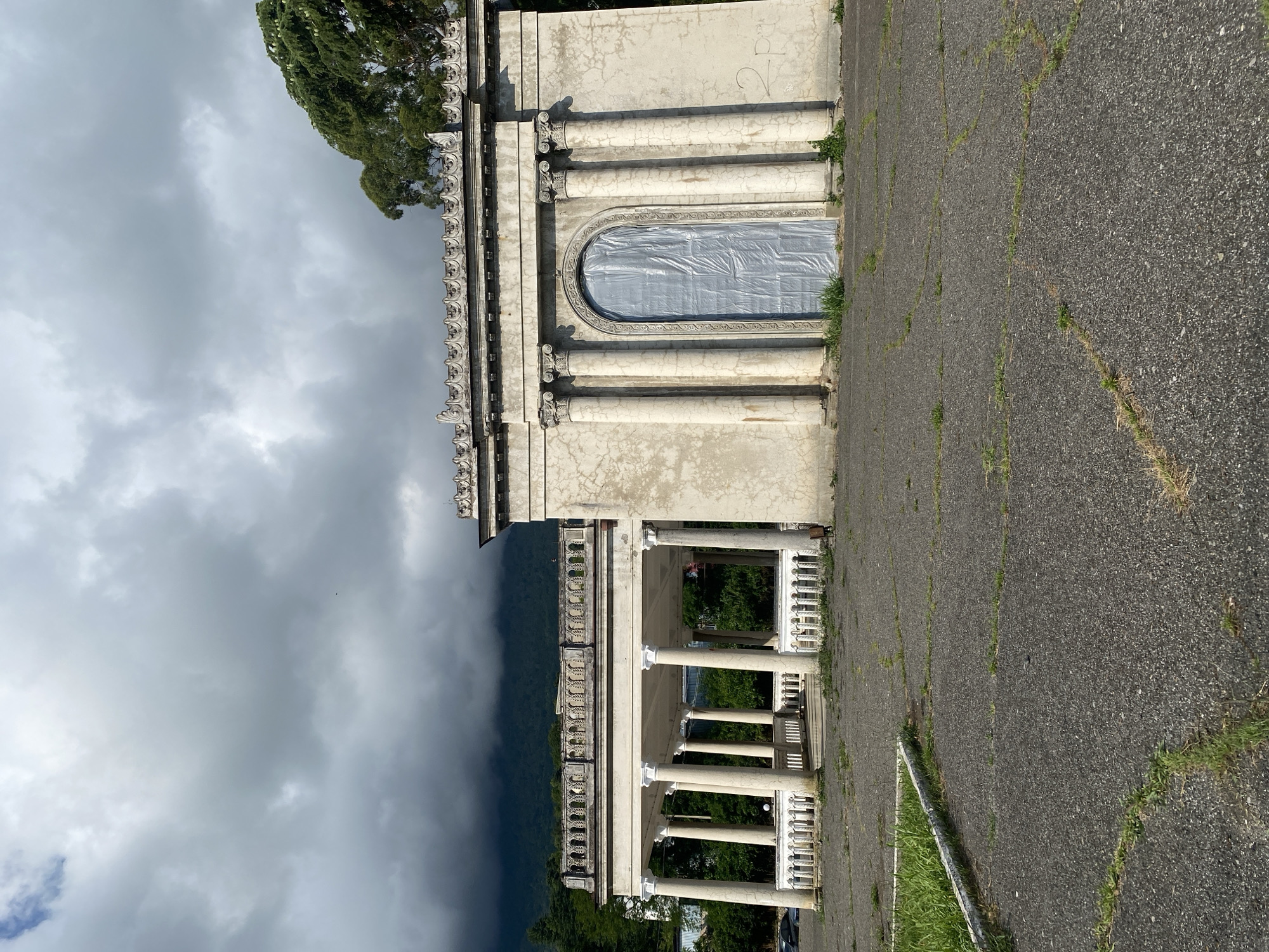 The image shows an abandoned building with classical architectural elements, possibly in Psakhara, Abkhazia. The building features a portico supported by Corinthian columns, a decorative balustrade, and a large arched entrance covered with a gray plastic tarp. The exterior walls are weathered and cracked, with some graffiti visible. The ground in front of the building is paved with asphalt that is broken and overgrown with grass. In the background, there are trees and a mountain shrouded in dark clouds. The sky is overcast with heavy gray clouds. There are no people or animals visible in the scene.