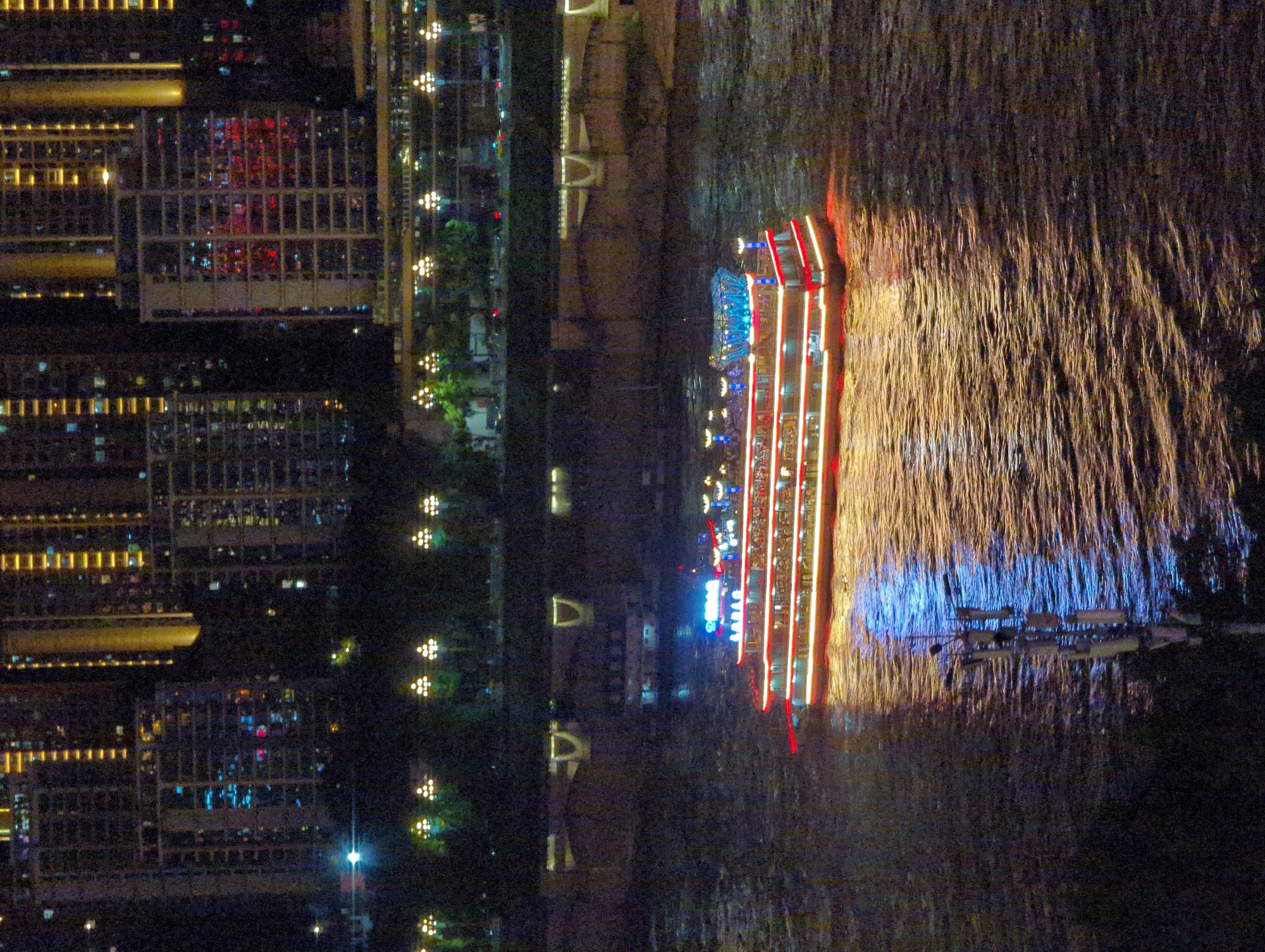 The image depicts a nighttime scene in Yuzhong District, China, featuring a brightly lit cruise ship on a river. The ship, with multiple decks adorned with red and white lights, is the central focus. Its illuminated reflection shimmers on the dark, rippling water. In the background, a cityscape of tall buildings rises, their windows glowing with a mixture of yellow, blue, and red lights, indicating a dense urban environment. A bridge structure is visible in the mid-ground, spanning the river. The overall impression is one of a vibrant, illuminated night scene in a major Chinese city.