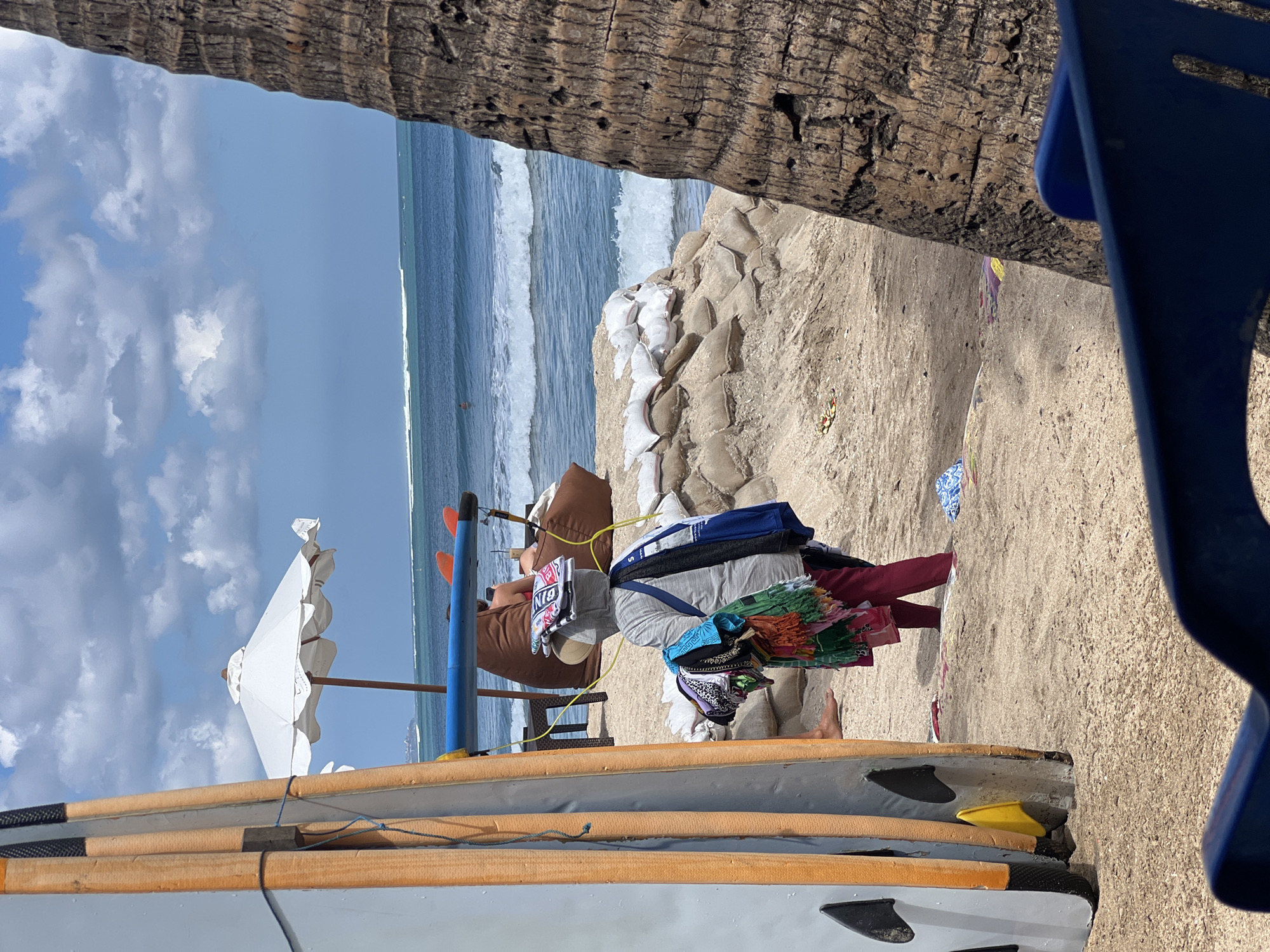 A beach scene in Kuta, Indonesia, under a partly cloudy sky. In the foreground, two surfboards are propped up on the sand. A person wearing a grey hoodie and maroon pants is carrying a stack of colorful cloths on their head and a blue bag over their shoulder. The person is standing on a sandy beach with several sandbags forming a barrier behind them. In the background, the ocean waves are breaking, and a white umbrella stands on the sand. A palm tree trunk is visible on the right side of the frame.