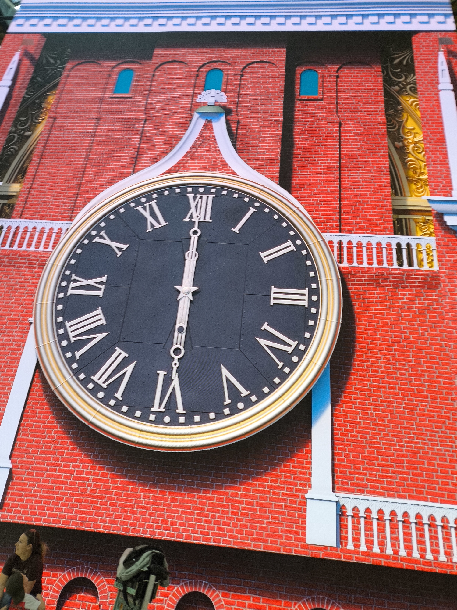 The image displays a large digital screen located in Khimki, Russia, depicting a stylized red brick clock tower. At the center of the screen is a prominent black clock face with white Roman numerals and white minute markers. The clock hands, also white and intricately designed with a star element and circular cutouts, are positioned vertically, indicating a time of approximately 6:00. The clock face is encased within a metallic gold and white bezel.
Above the clock, the red brick facade features two small arched, light-blue windows on the left and one on the right. A decorative white architectural element, resembling a stylized tree, sits directly above the clock, supported by white curved structures that emerge from the clock's upper edge. To the right of the clock, a white balustrade with multiple vertical supports extends horizontally, partially obscuring a gold, ornate architectural detail with swirling patterns. A slender white spire-like structure is also visible on the far right. Similar, less distinct architectural elements are present on the far left. The brick pattern of the facade appears uniform with some subtle shading.
In the lower left corner of the image, two individuals are partially visible. One person, with dark hair, is seen from the chest up, wearing a dark t-shirt and facing left. Another person, mostly obscured, is to their right. Adjacent to these individuals, a dark green or gray stroller or bag with wheels is present. The bottom edge of the brick facade shows multiple arched recesses. The lighting suggests an external light source, casting shadows to the right of the white architectural features and below the clock. The entire scene is vibrant, consistent with a large-scale illuminated display.