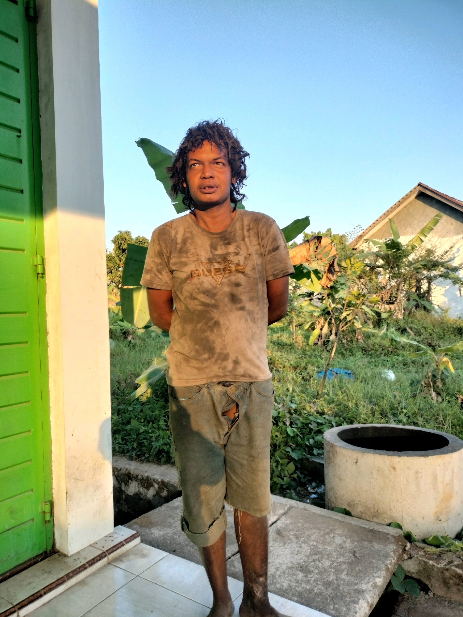 A man with curly, dark hair and an orange complexion stands in front of a white wall and a green door, with his hands clasped behind his back. He is wearing a dirt-stained t-shirt with the word "GUESS" visible on it, and ripped denim shorts. The background shows lush green vegetation, a house with a triangular roof, and a concrete well. The scene is bathed in the warm light of what appears to be late afternoon or early morning. The man's attire and the rustic surroundings suggest a location in a rural or semi-rural area. The image is set in Tersono, Indonesia.