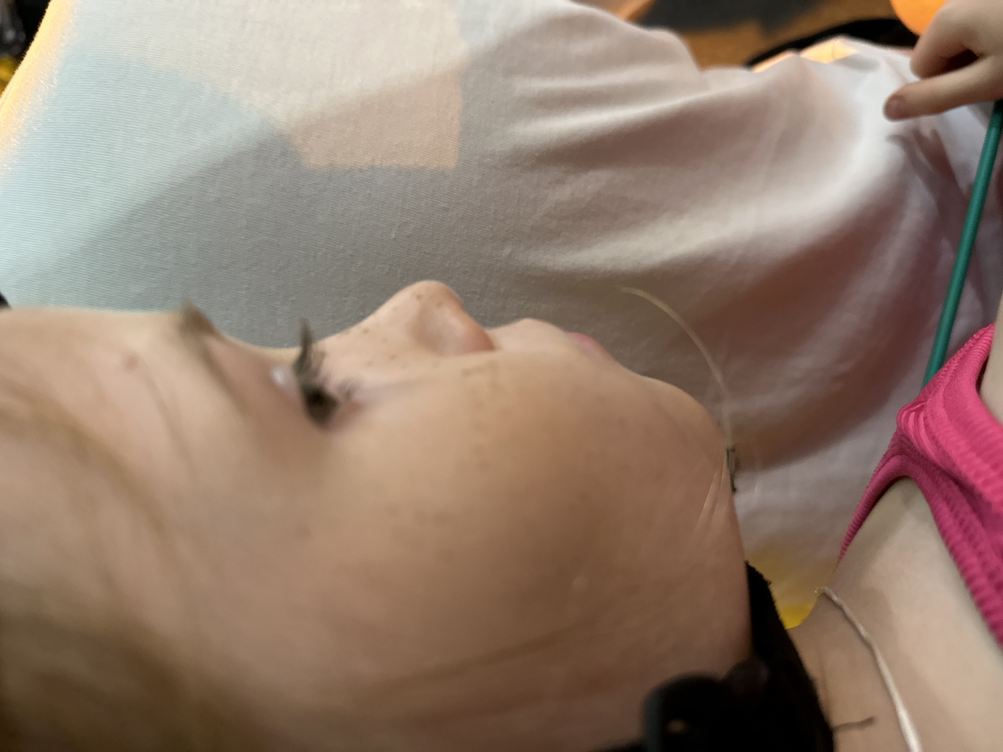 A close-up, profile view captures the left side of a young girl's face, showing her blonde hair, light skin with freckles, and a green necklace. She wears a bright pink tank top. Her eyes are looking down and to the left. Her head is turned to the right, and her face is slightly blurred. Behind her, a light blue, textured fabric is visible, likely clothing or a blanket. The lighting is dim, with warm tones suggesting an indoor setting, possibly in Nha Trang Ward, Vietnam.