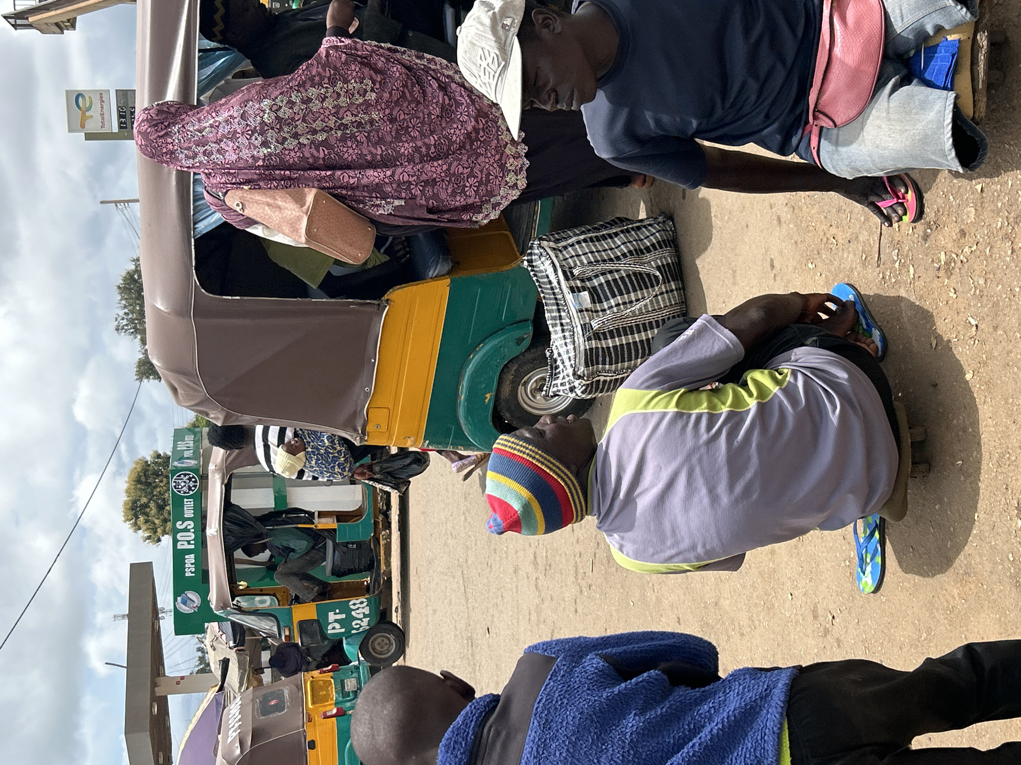 The image depicts a street scene in Bar Jirgi Summa, Nigeria, with a focus on several auto-rickshaws, commonly known as "keke napeps." In the foreground, two men are seated on what appear to be improvised wooden stools or seats on the ground. One man is wearing a blue fleece jacket and black pants, his back to the camera. The other man, facing to the right, is wearing a striped beanie, a grey shirt with a lime green trim, and dark pants. He is sitting on a flat board, and a pair of colorful flip-flops are on the ground near his feet.
To the right of the foreground figures, a third man is visible, wearing a dark blue t-shirt, light blue jeans, and a light-colored baseball cap. He is also seated, with a pink fanny pack around his waist. His hands are visible, with one resting near his knee, and he is wearing bright pink flip-flops.
Behind these men are two auto-rickshaws. The one on the left is predominantly green with yellow accents and has the license plate "PT 6248" clearly visible. A person is seated inside, partially obscured. The auto-rickshaw on the right is also green and yellow, with a brown canopy. A woman wearing a vibrant purple and pink patterned headscarf and a light pink bag slung over her shoulder is standing next to this auto-rickshaw, partially entering or exiting. Another person in a black garment is seated inside this vehicle. A large black and white checkered duffel bag is placed on the ground next to the right auto-rickshaw.
In the background, a gas station structure with a canopy and a "TotalEnergies" sign is visible. The ground surface appears to be unpaved, with loose dirt and sand. The sky is overcast with some visible clouds. The overall impression is of a busy roadside location, likely a transport hub or market area.