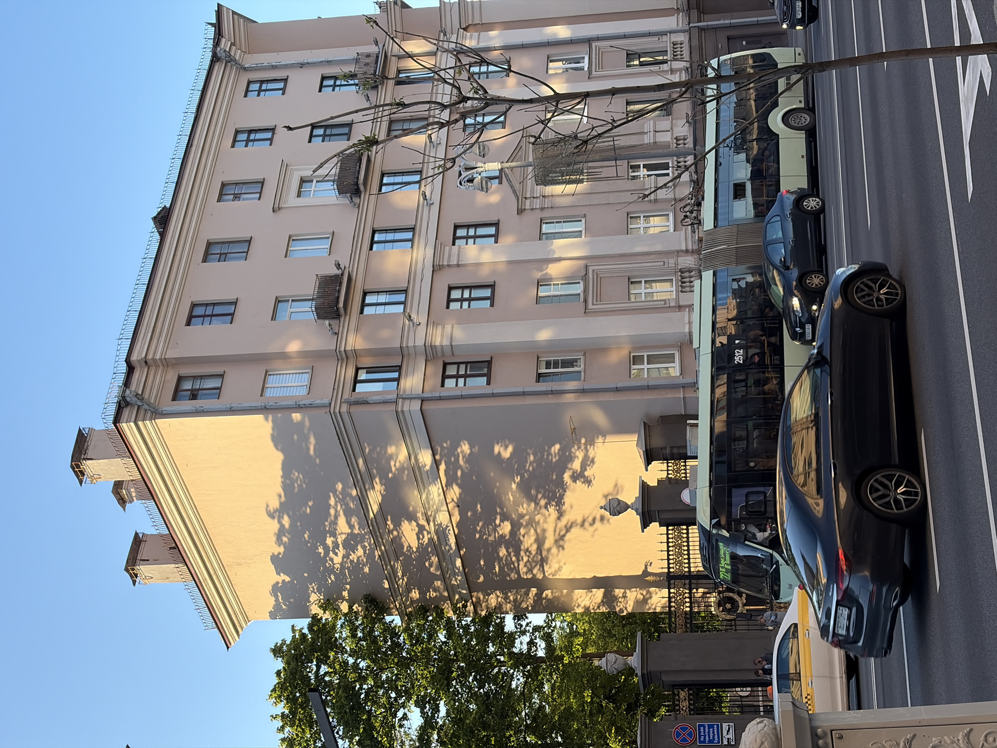 The image captures an urban scene in Minsk, Belarus, featuring a tall, light-colored building with multiple windows, some with small balconies. A tree casts shadows on the building's facade. Alongside the building, there's a street with a bus and several cars. A decorative metal fence with pillars and what appears to be a no-parking sign is visible at the street's edge. The sky is clear and blue. The image is taken from a slightly low angle, looking up at the building.