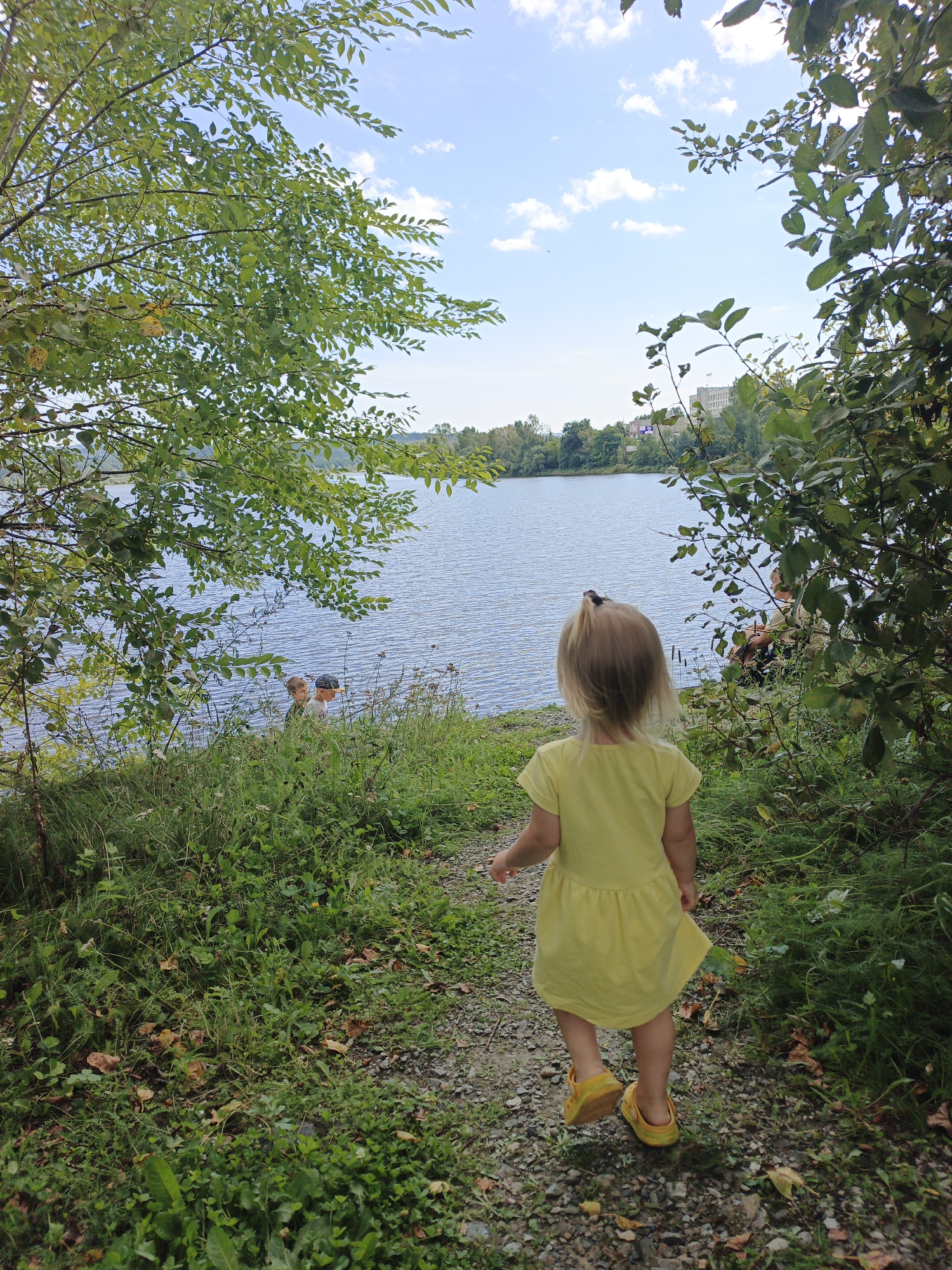 In Zlatoust, Russia, a young girl with blonde hair in a yellow dress and yellow shoes walks away from the camera on a gravel path. She is heading towards a large body of water, a lake or river, which reflects the blue sky and white clouds. Green trees and foliage surround the path and the water's edge. Two children, possibly boys, are visible near the water's edge on the left side of the frame, partially obscured by grass. In the distance across the water, there are trees and buildings, suggesting the presence of a town or city. The scene appears to be an outdoor recreational area.