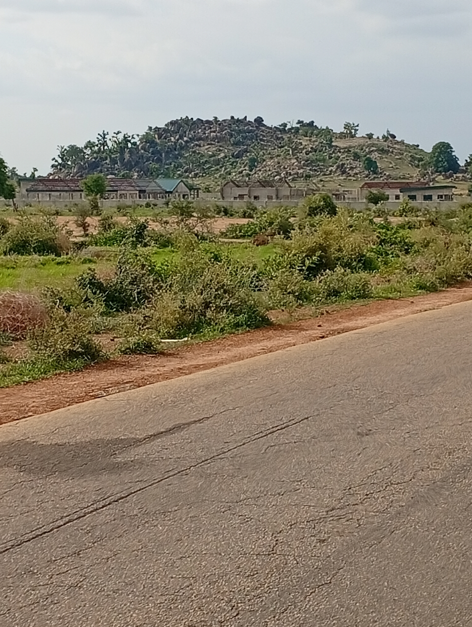 The image shows a rural landscape with a road in the foreground and a rocky hillside with buildings in the background. The road is paved and appears to be in fair condition, with some cracks and markings. On the side of the road, there is a dirt embankment covered with vegetation. In the mid-ground, there are several buildings, some of which appear to be under construction. These buildings are low-lying and rectangular, with various roof styles, including some with green roofs. The buildings are situated in a vegetated area with trees and shrubs. The background features a prominent rocky hill dotted with trees and some sparse vegetation. The sky is overcast with a light gray tone. The location is identified as Shuwa, Nigeria.