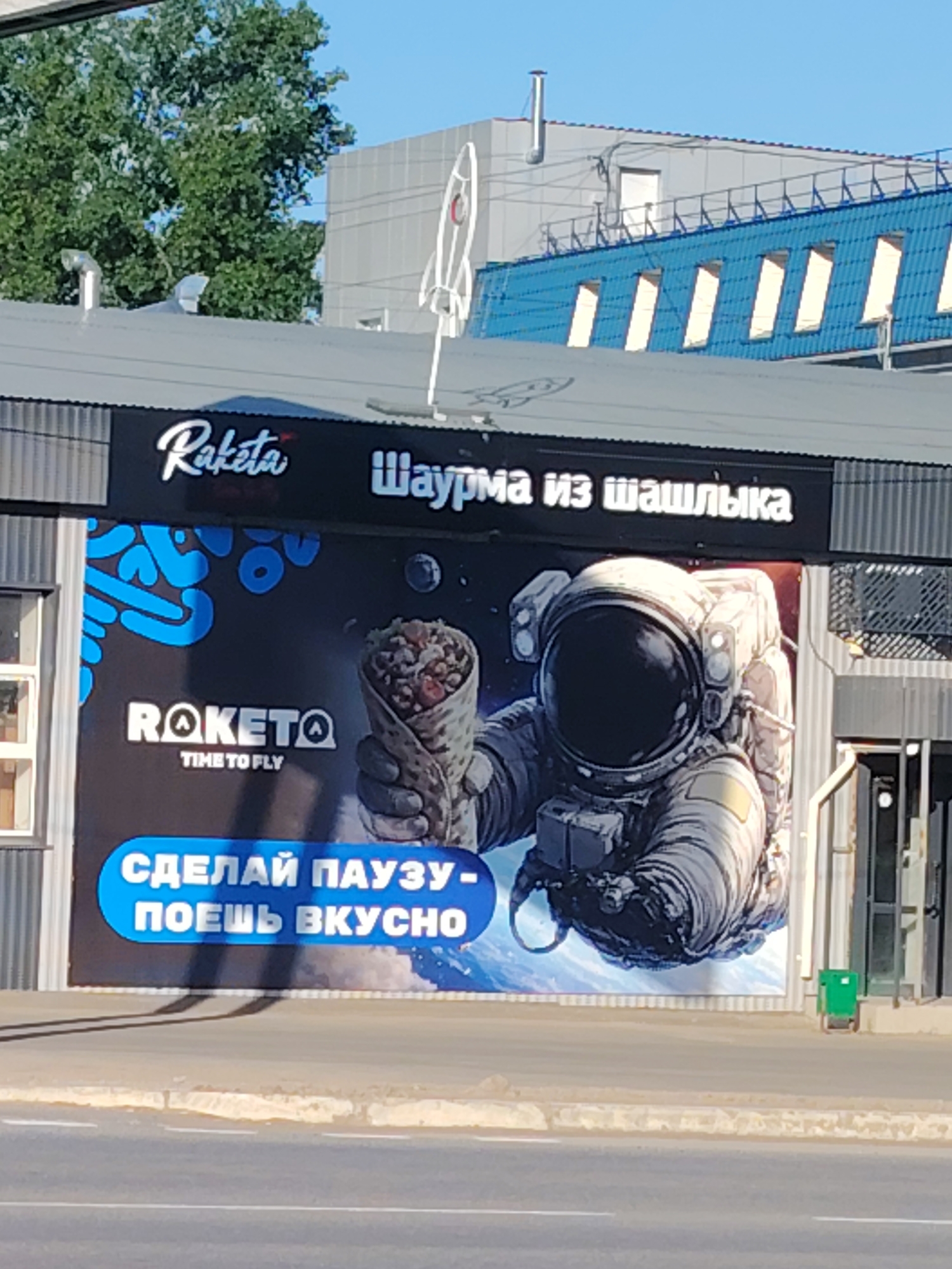 A photograph captures a commercial building featuring a large advertisement for "Raketa" (rocket in Russian), located in the city of Chaikovsky, Russia. The sign above displays the restaurant's name, "Raketa," alongside the phrase "Shawarma iz shashlyka" (shawarma from shashlik). The billboard below showcases an astronaut holding a shawarma against a backdrop resembling Earth. Below the astronaut and the shawarma, the text "Sdelay pauzu - poes vkusno" (take a break - eat tasty) is displayed in a bright blue banner. The building itself has a gray roof with a decorative rocket on top. Behind the building are trees and a building with a blue roof. The image is taken from street level, showing a segment of a road in the foreground.