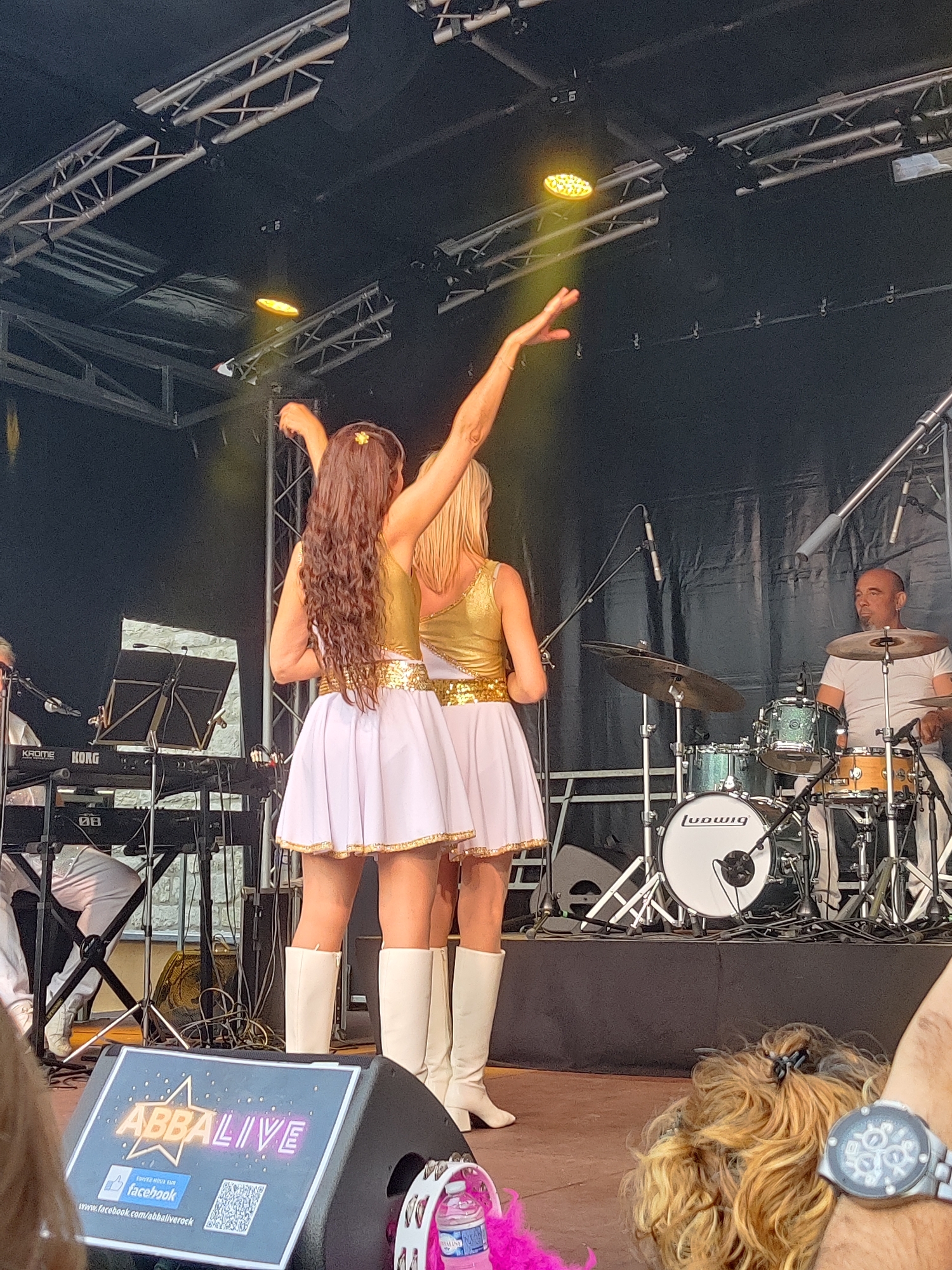 The image depicts an outdoor concert stage in Marche-en-Famenne, Belgium. Two female performers, dressed in gold sequined tops and white A-line skirts with gold trim, stand facing away from the camera, with their arms raised. The performer on the left has long, dark, wavy hair adorned with a small flower. The performer on the right has blonde hair. To the left of the performers, a keyboard player is seated behind a KORG keyboard. To the right, a male drummer is positioned behind a drum kit, including cymbals and bass drum, which has the "Ludwig" brand visible. Overhead, stage lighting rigs and trusses are visible against a dark background. In the foreground, a speaker with an "ABBA LIVE" logo is prominent, displaying a Facebook link and a QR code. A person's arm, wearing a wristwatch, is visible in the lower right corner. The overall environment suggests a live performance event.