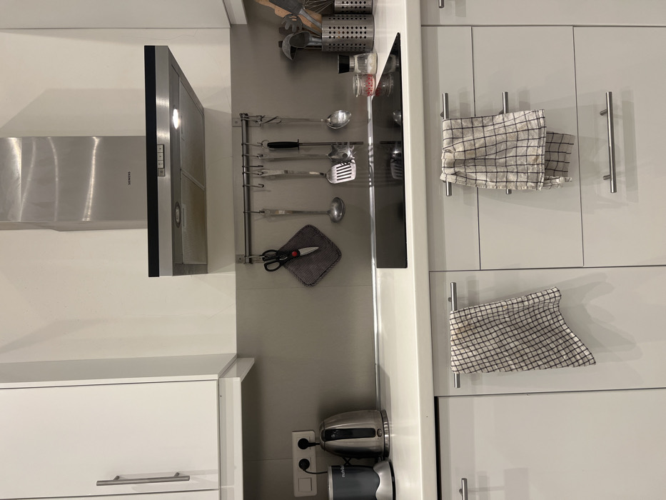 A narrow kitchen view showcasing white cabinets. Cooking utensils hang on a rack above a black-handled scissor on a gray square pot holder. Two dish towels hang from metal bars on the adjacent cabinet doors. A stainless steel and black range hood is above a lower white cabinet. A coffee maker sits on the counter near an electrical outlet.