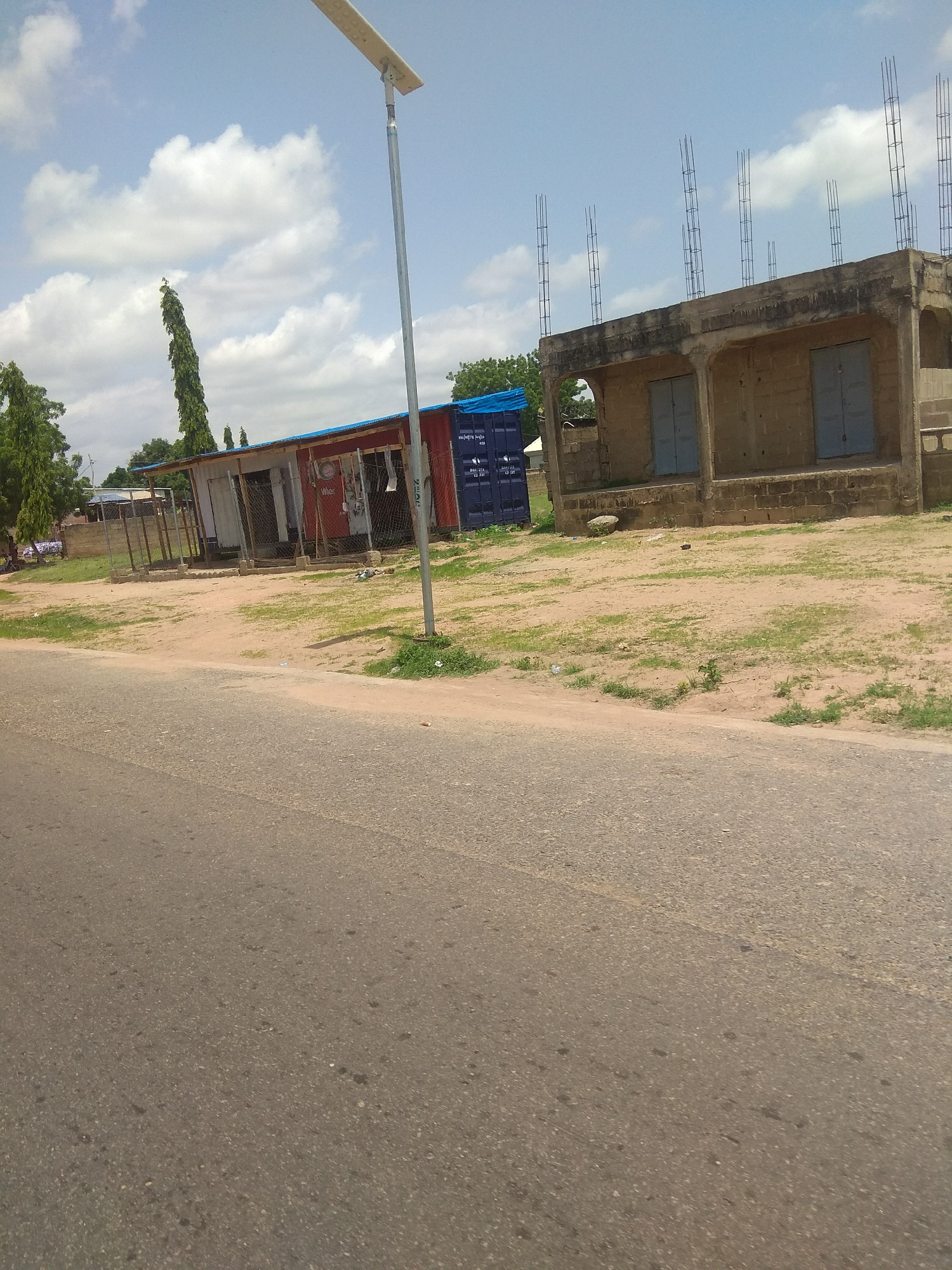 This media file depicts an outdoor scene in Jalingo, Nigeria. The subjects include several buildings in varying states of construction and completion, a road in the foreground, and a prominent solar-powered street light on a tall metal pole.
On the left, there is a row of small, seemingly commercial structures with red corrugated metal roofs. One of these structures appears to be a shop with a white facade and a visible chain-link fence. Next to it, a blue shipping container with some visible text is parked. Further to the left, there is a fenced area, possibly a yard or part of a property.
To the right of the shops, a more substantial building is under construction. It has a concrete structure with visible rebar extending from the top, indicating it is in the process of having an upper floor added. This building has a series of arched openings on the ground floor, with two large blue-paneled doors visible. The walls of this building appear to be made of stone or concrete blocks.
The ground in front of the buildings is a mixture of sandy soil and patchy green grass. A paved road runs across the bottom of the frame. In the background, several tall, slender green trees are visible.
The weather appears to be partly cloudy with a bright sky, suggesting it is daytime. The sun's glare on the road and the shadows cast indicate it is likely midday. There are no people visible in the frame. The overall impression is one of a developing area, with both existing small businesses and new construction projects.
Visible text includes markings on the blue shipping container, though it is too small and blurry to decipher specific details. The solar panel on the street light is also a notable detail.