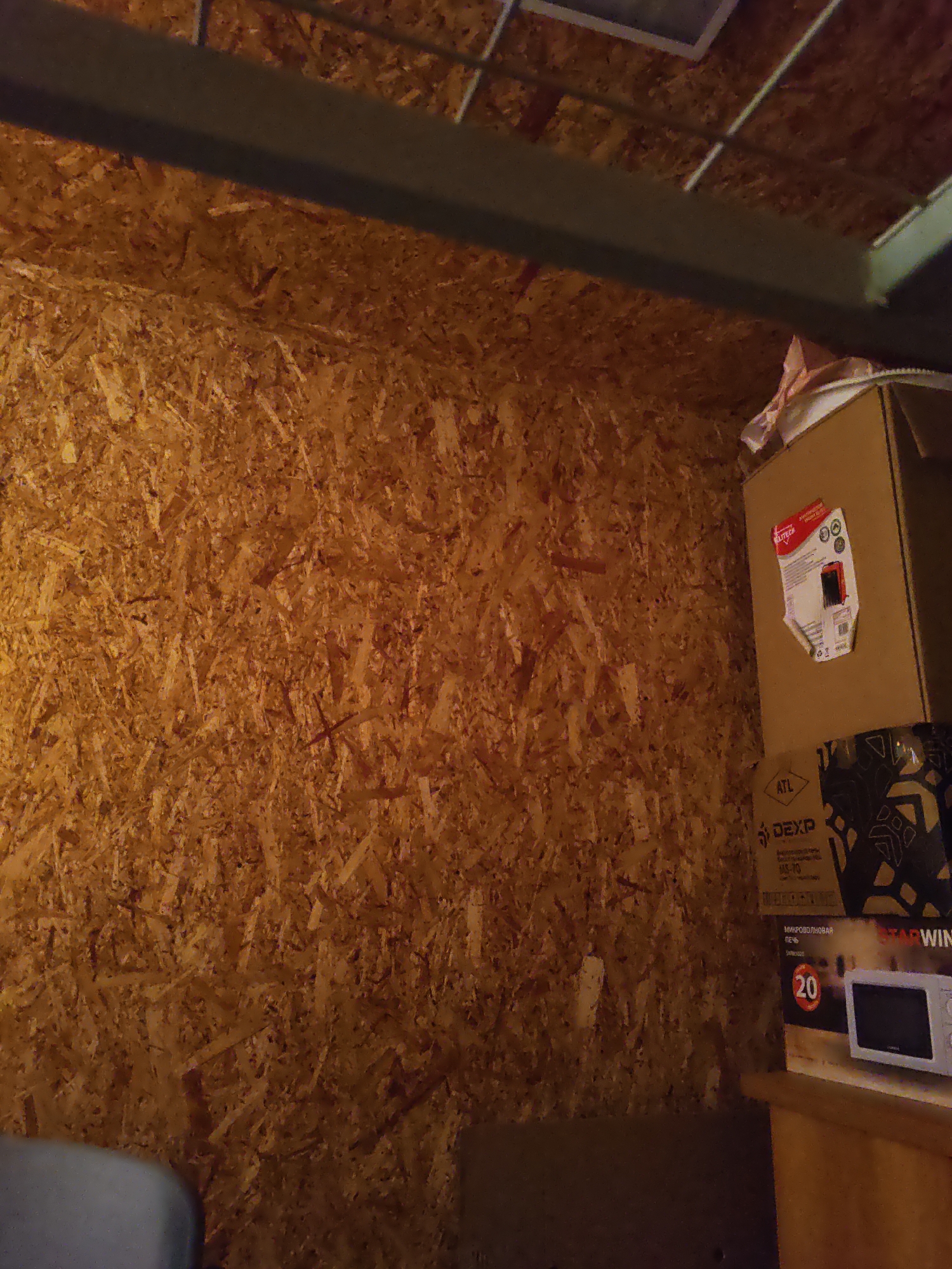 The image depicts the interior of what appears to be a storage space or a closet. The walls are constructed of OSB (Oriented Strand Board), a wood-based panel product. In the background, there are several cardboard boxes stacked, with some labels partially visible. One box on the right has a red and white label with the text "ELITECH" and some circular icons. Below this, another box is visible with the text "DEXP" and "ATL" on it. Further down, a microwave oven is visible on a wooden shelf, with a circular label showing the number "20" and the Russian word "ПЕЧЬ" (pech), meaning oven. The overall lighting suggests an indoor environment, likely with artificial lighting. The specific location of Платошино, Russia, cannot be determined from the visual information provided in the image itself, as there are no discernible landmarks or contextual clues to indicate this geographical location.
