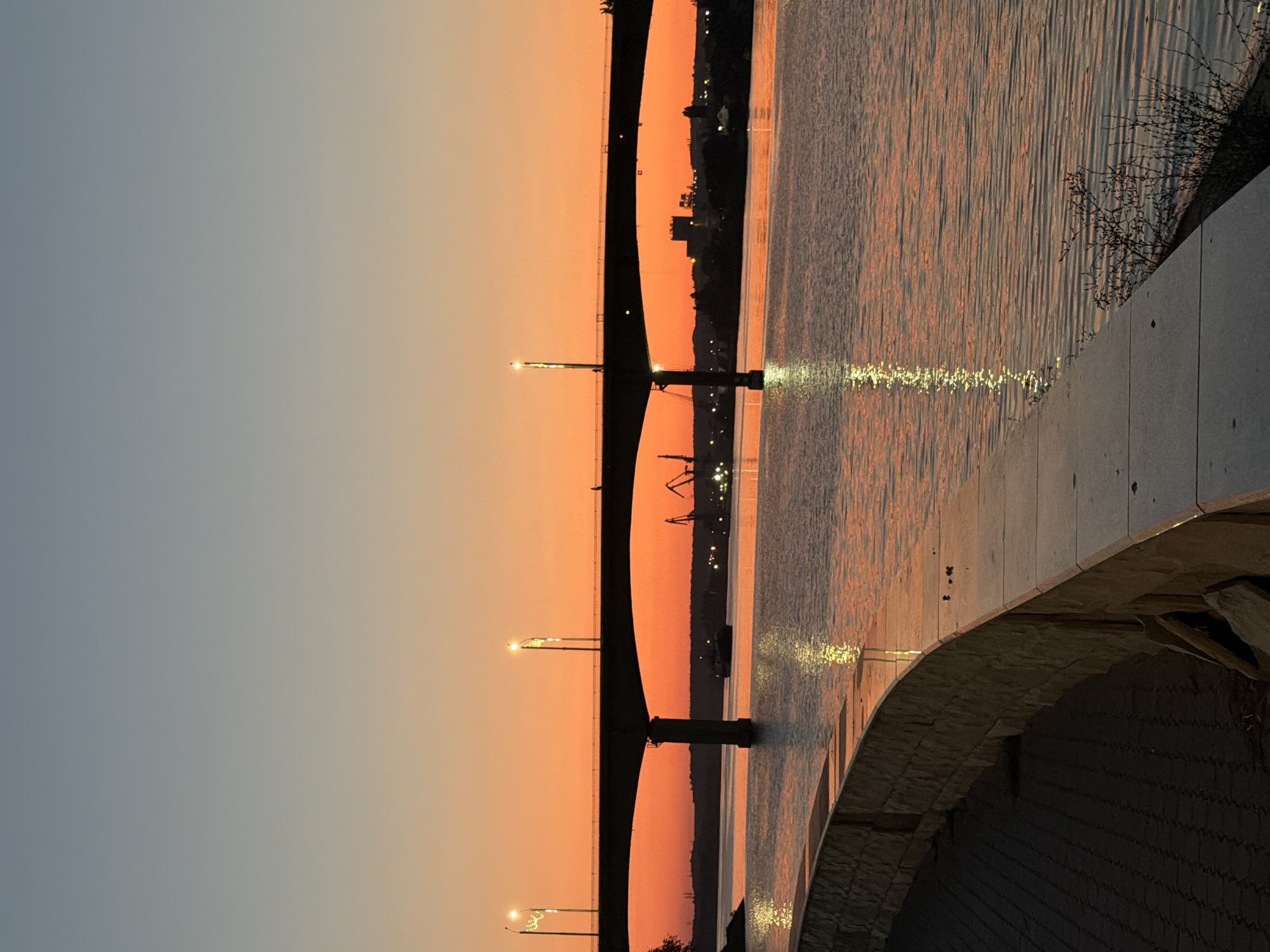The image depicts a sunset over the Volga River in Kamyshin, Russia, viewed from a promenade. A wide, concrete bridge spans the river, silhouetted against the orange and blue gradient of the twilight sky. Several streetlights on the bridge are illuminated. In the distance, industrial cranes and buildings are visible on the far bank. The river's surface reflects the warm colors of the sunset, with streaks of light from the bridge's lamps shimmering on the water. A paved promenade with a stone retaining wall curves along the foreground, leading the eye towards the river and the bridge. The scene is peaceful, with no people or animals clearly visible, save for a lone figure on the bridge.