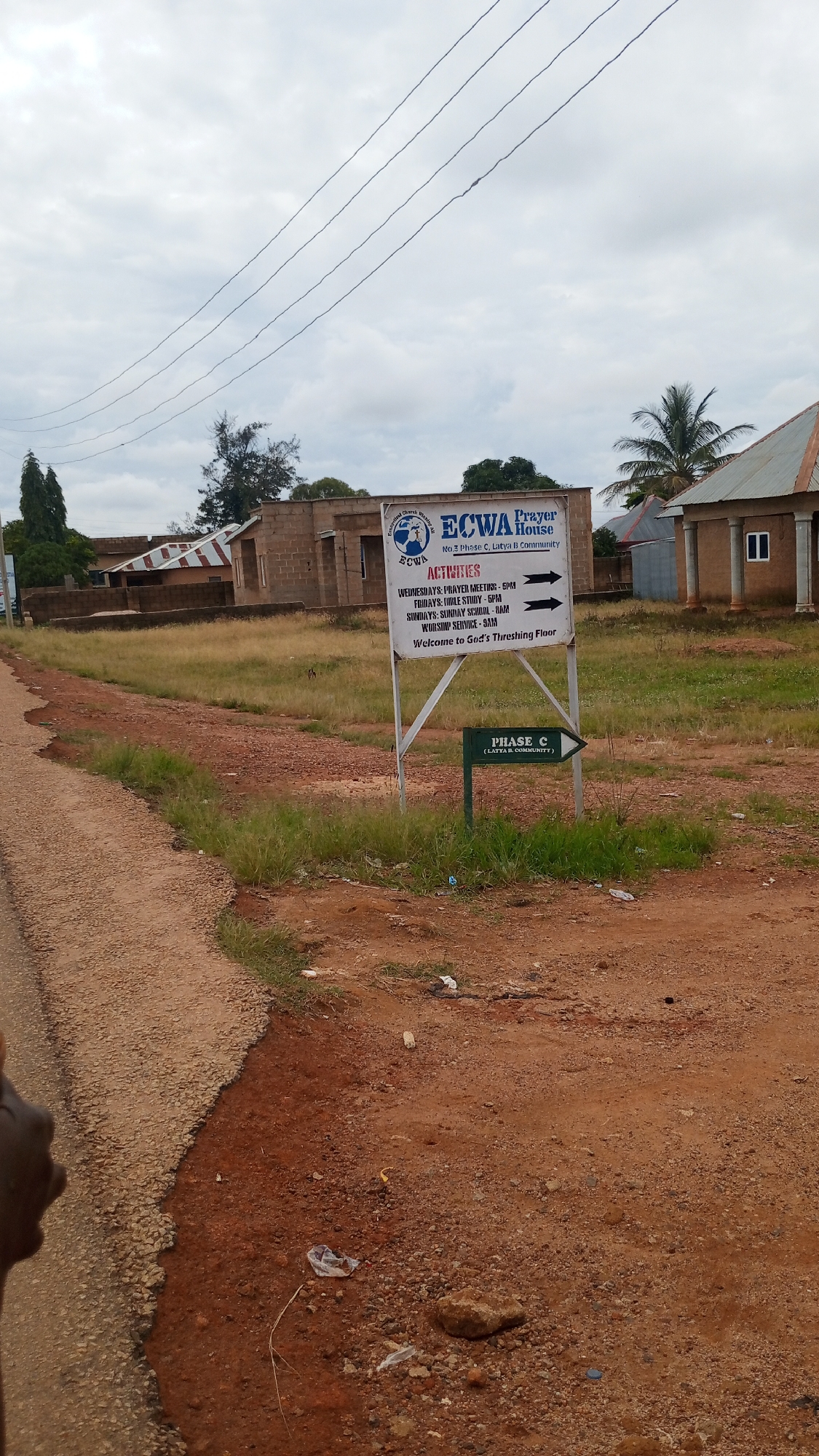 A slightly overcast, cloudy day is captured in this medium shot of a dirt road with a signpost in front of buildings. The signpost is white with black lettering. At the top, it says "ECWA Prayer House". Below that is "No 3 Phase C, Latya fi Community". Underneath that is the word "ACTIVITIES" in red, followed by a list of activities and their times, each with a black arrow pointing to the right. The last line of text on the sign is "Welcome to God's Threshing Floor". Below this main sign is a smaller green sign with white text that reads "PHASE C (LATYA B. COMMUNITY)" with a white arrow pointing to the right. Behind the signpost, there are several buildings constructed from brick and corrugated metal roofs. The ground in the foreground is dirt with some dry grass and scattered debris, including plastic bags and rocks. To the left of the frame, a small portion of a person's dark hand is visible at the edge of the road.