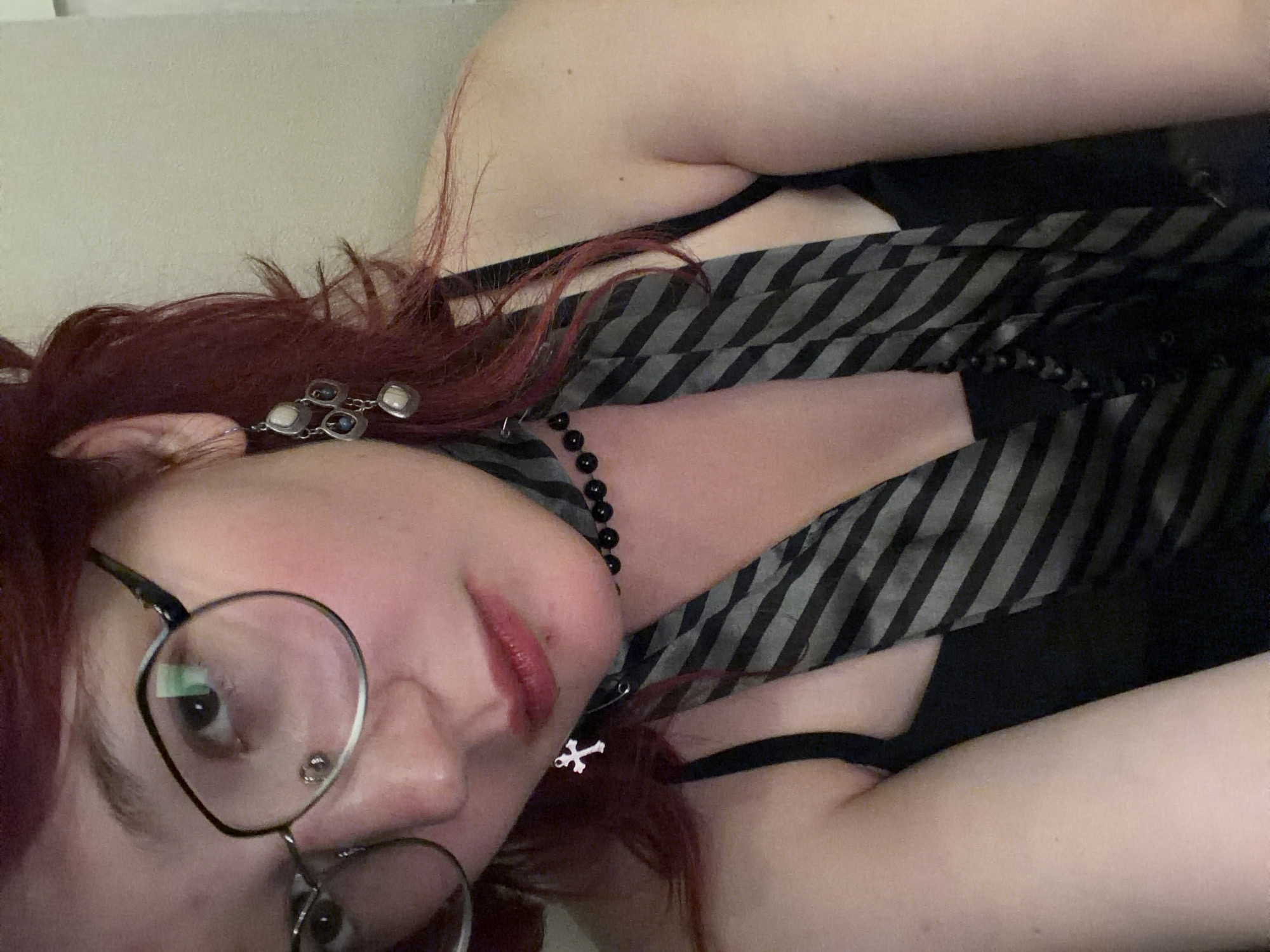 The image is a close-up, selfie-style photograph of a person with reddish-purple hair, wearing glasses and jewelry. The person is looking towards the camera, with their face slightly tilted. They are wearing a black top, a striped tie, a beaded necklace, and earrings. A small cross pendant is visible on their neck. The background is plain and light-colored, suggesting an indoor setting. There is no visible indication of the location being in Chelyabinsk, Russia.