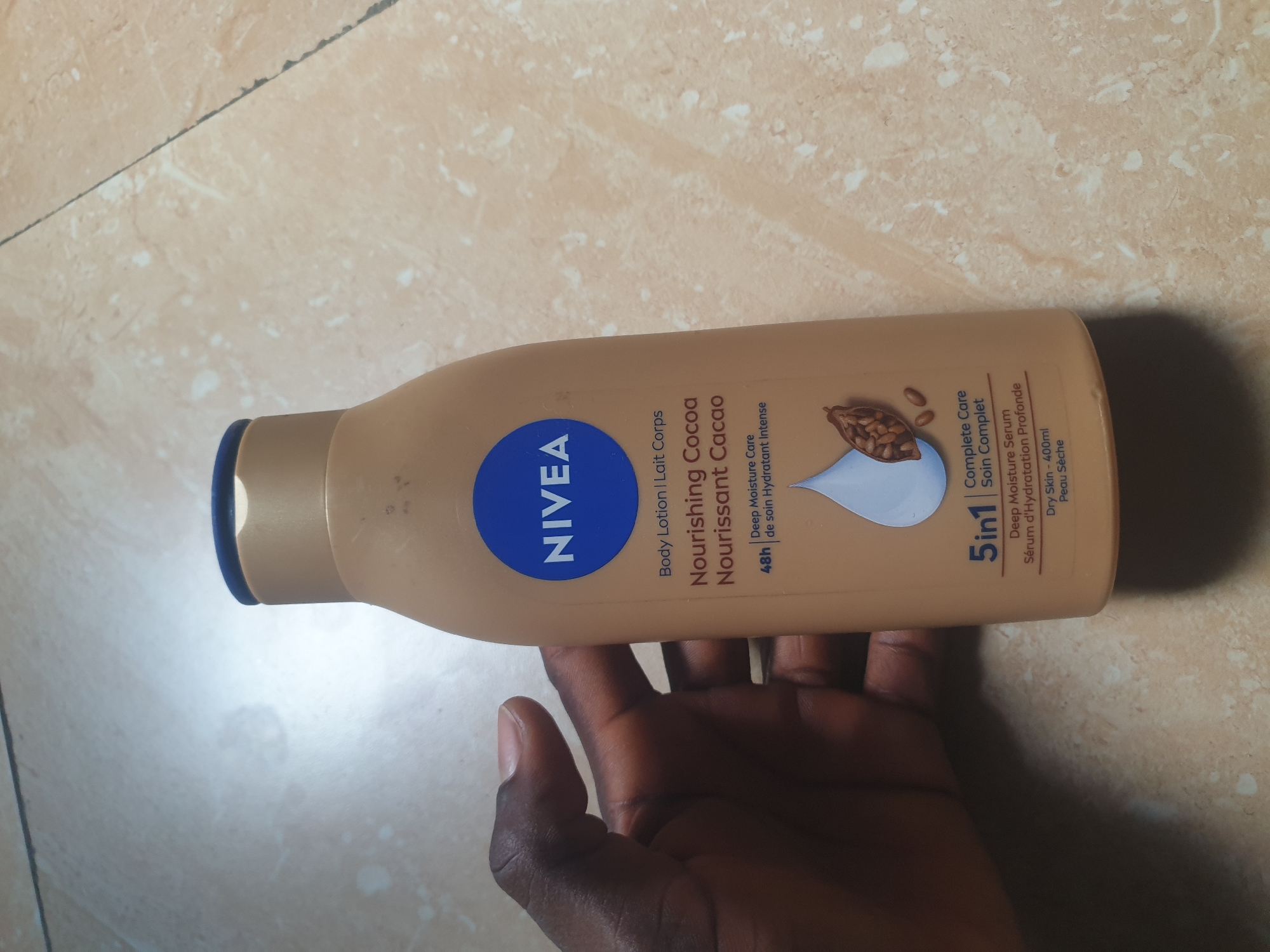 A Nivea Nourishing Cocoa Body Lotion bottle, 400ml, for dry skin, is depicted in a close-up. The light brown plastic bottle with a dark blue flip-top cap features the Nivea logo and product details in English and French, along with an illustration of a white droplet and cocoa beans. A dark-skinned hand holds the bottle from the left side, with fingers partially visible on the front surface. The bottle rests on a light-colored, patterned tiled floor with visible grout lines and faint smudges. The scene is located in the city of Jalingo, Nigeria.