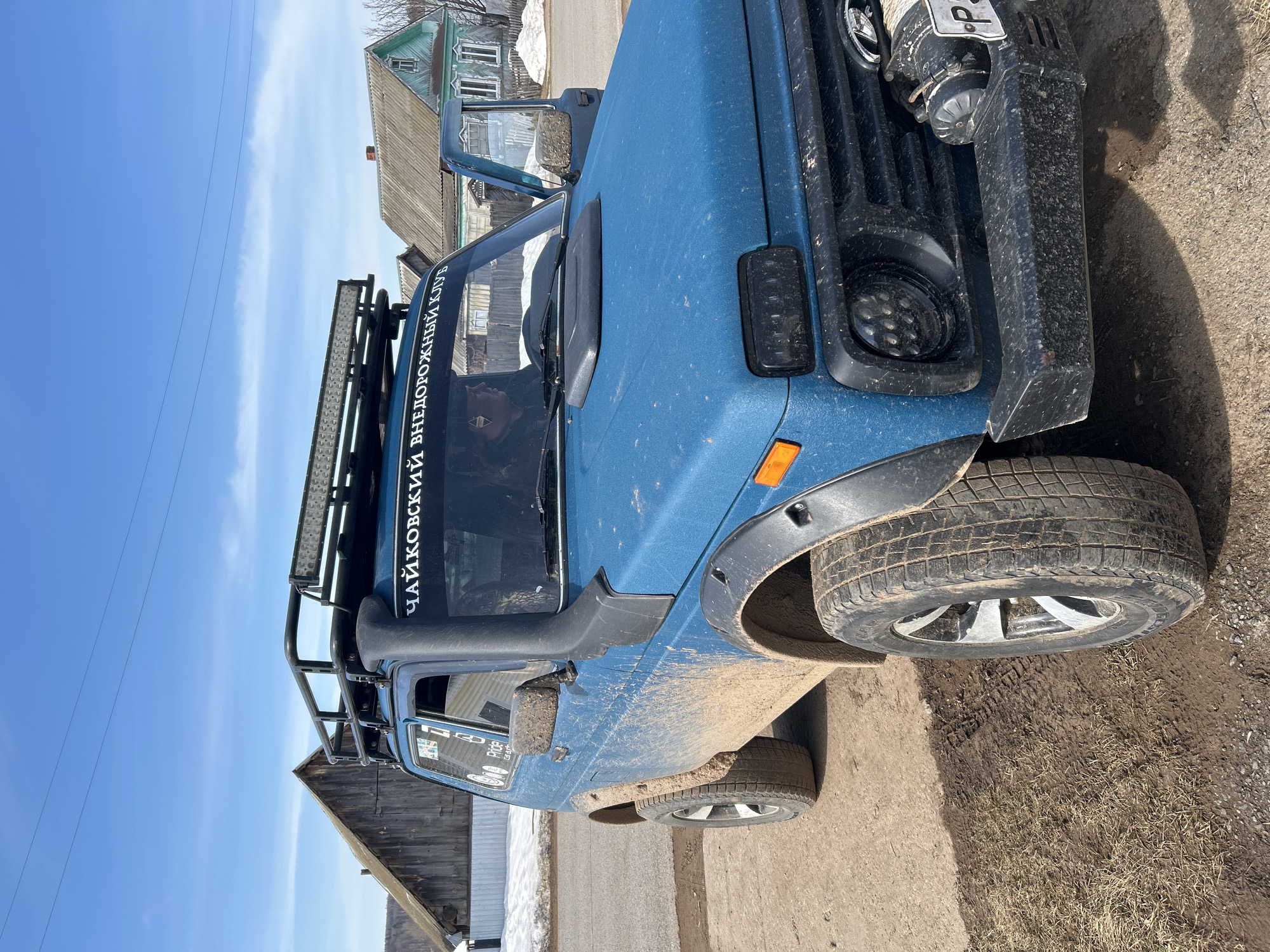 The image shows a blue off-road vehicle parked outdoors in the city of Уральское, Russia. The vehicle is covered in mud, suggesting it has recently been driven off-road. It has a black roof rack with an LED light bar. The windshield has a sticker with white lettering that reads "ЧАЙКОВСКИЙ ВНЕДОРОЖНЫЙ КЛУБ". The vehicle has aftermarket headlights, a snorkel intake on the driver's side, and a winch mounted on the front bumper. The background includes a wooden house, a dirt road, and a blue sky. There is some snow still visible on the ground near one of the houses.