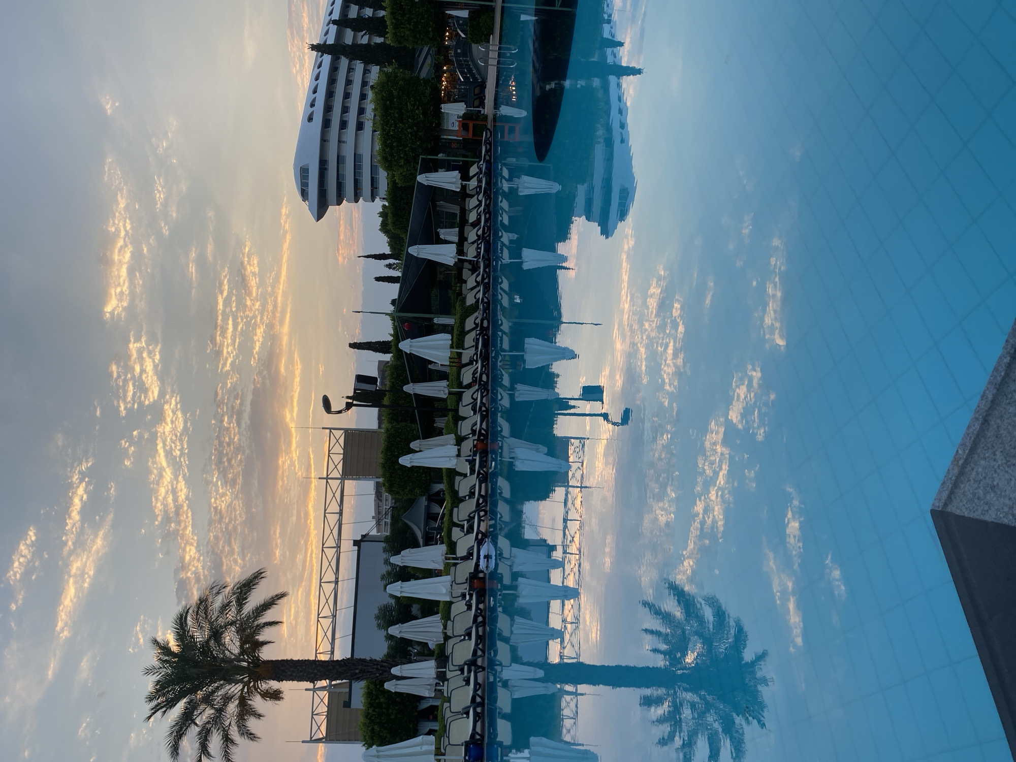 The image depicts a swimming pool at a resort in Aksu, Turkey, during a sunset. The sky is filled with scattered clouds tinged with orange and yellow hues. A palm tree stands tall on the left side of the frame, its reflection visible in the still water of the pool. Along the edge of the pool, rows of closed white umbrellas and empty lounge chairs are arranged. In the background, a modern hotel with a distinctive curved white facade is visible, along with cypress trees and other greenery. The water of the pool is a vibrant blue, reflecting the sky and surrounding elements.