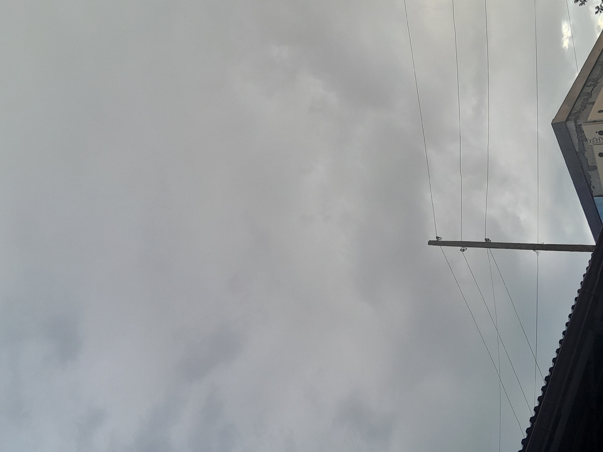 The image shows a view looking up at an overcast sky. Several power lines stretch horizontally across the frame, supported by a wooden utility pole. In the upper right corner, a portion of a building is visible, with its angled roofline and what appears to be construction materials on its side. The sky is a uniform grey, suggesting a cloudy or possibly stormy day. There are no people visible in the image, and no specific activity is taking place. The scene is urban or suburban, indicated by the presence of power lines and a building. The overall mood is neutral, with the overcast sky creating a somewhat somber or subdued atmosphere. No text is clearly legible in the image.